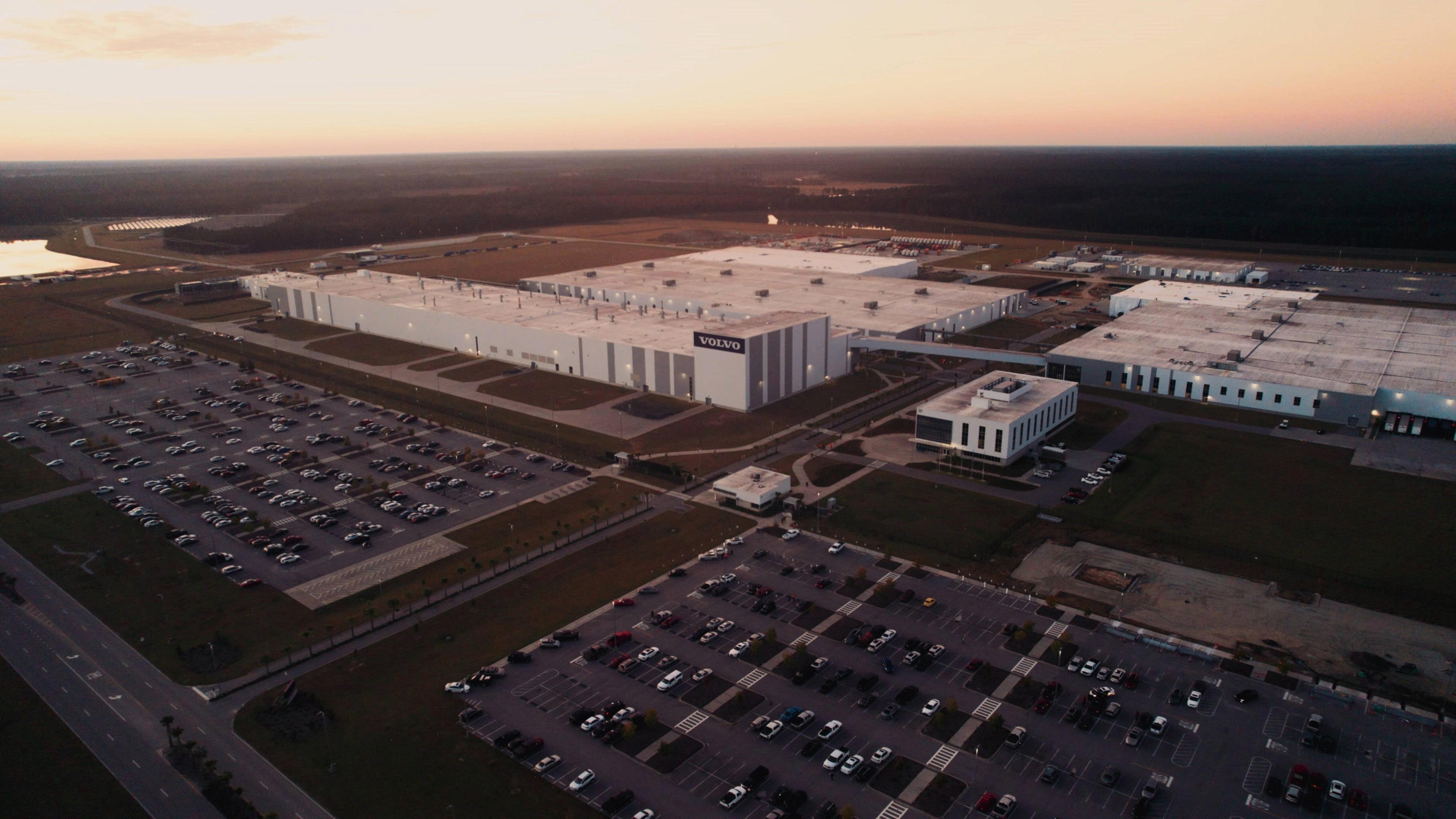 Area industrial da fábrica da Volvo em Charleston, Carolina do Sul, tirada do alto. O céu, em tons de azul, laranja e amarelo, sugere o nascer ou o pôr do sol, e a paisagem é dominada por grandes edifícios fabris modernos, predominantemente brancos e cinzentos, com o logótipo da Volvo bem visível no centro. Os vastos parques de estacionamento cheios de veículos no primeiro plano e a estrada que os delimita destacam a escala da operação, enquanto o horizonte é marcado por uma vasta extensão de floresta escura, que contrasta com o ambiente industrial.
