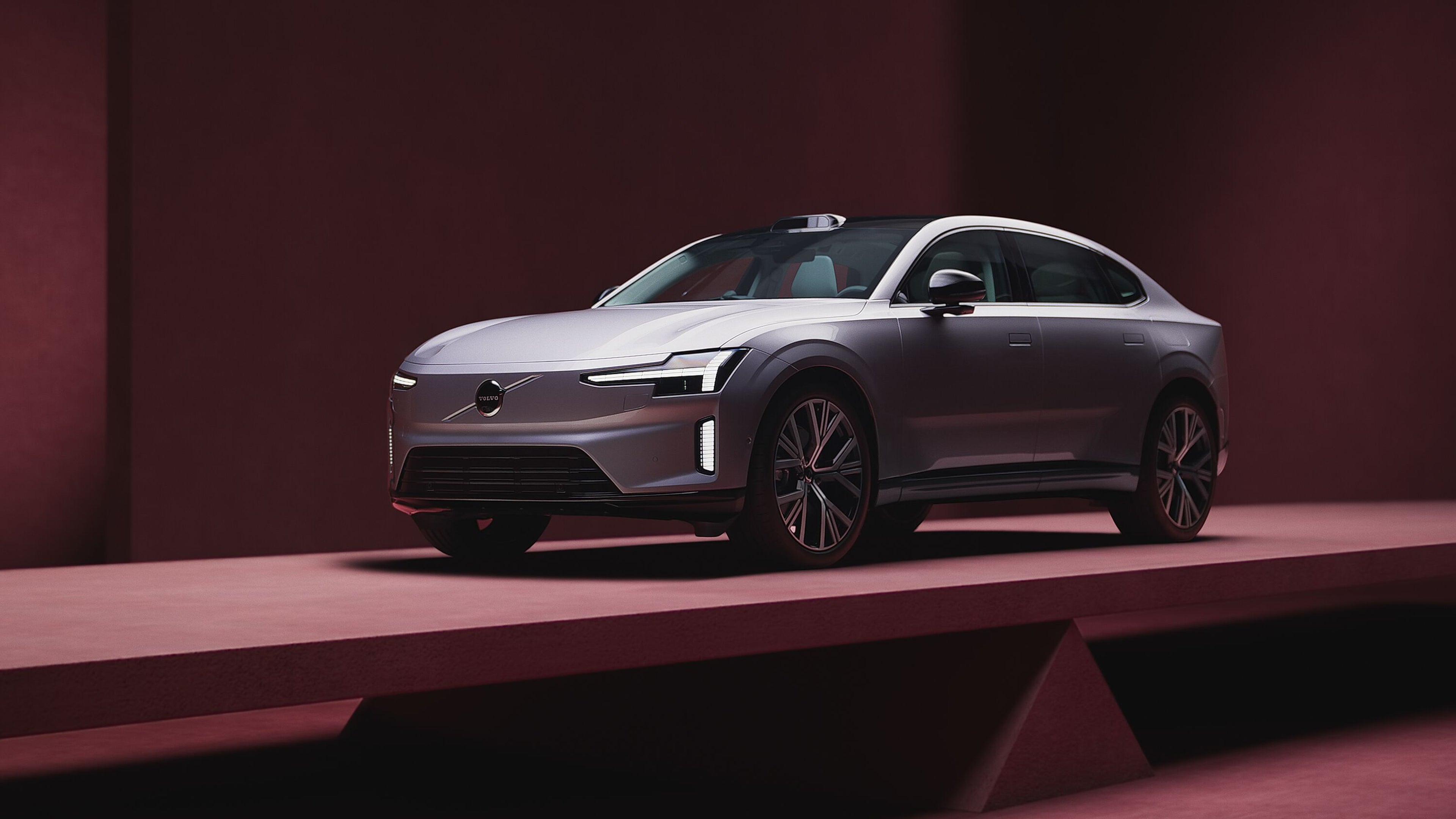 Поглед од преден агол на Volvo ES90, нашиот следен целосно електричен автомобил на кој ќе биде изложен, со модерни LED фарови и аеродинамичен дизајн, прикажан во минималистичко студио со длабоко црвено осветлување.