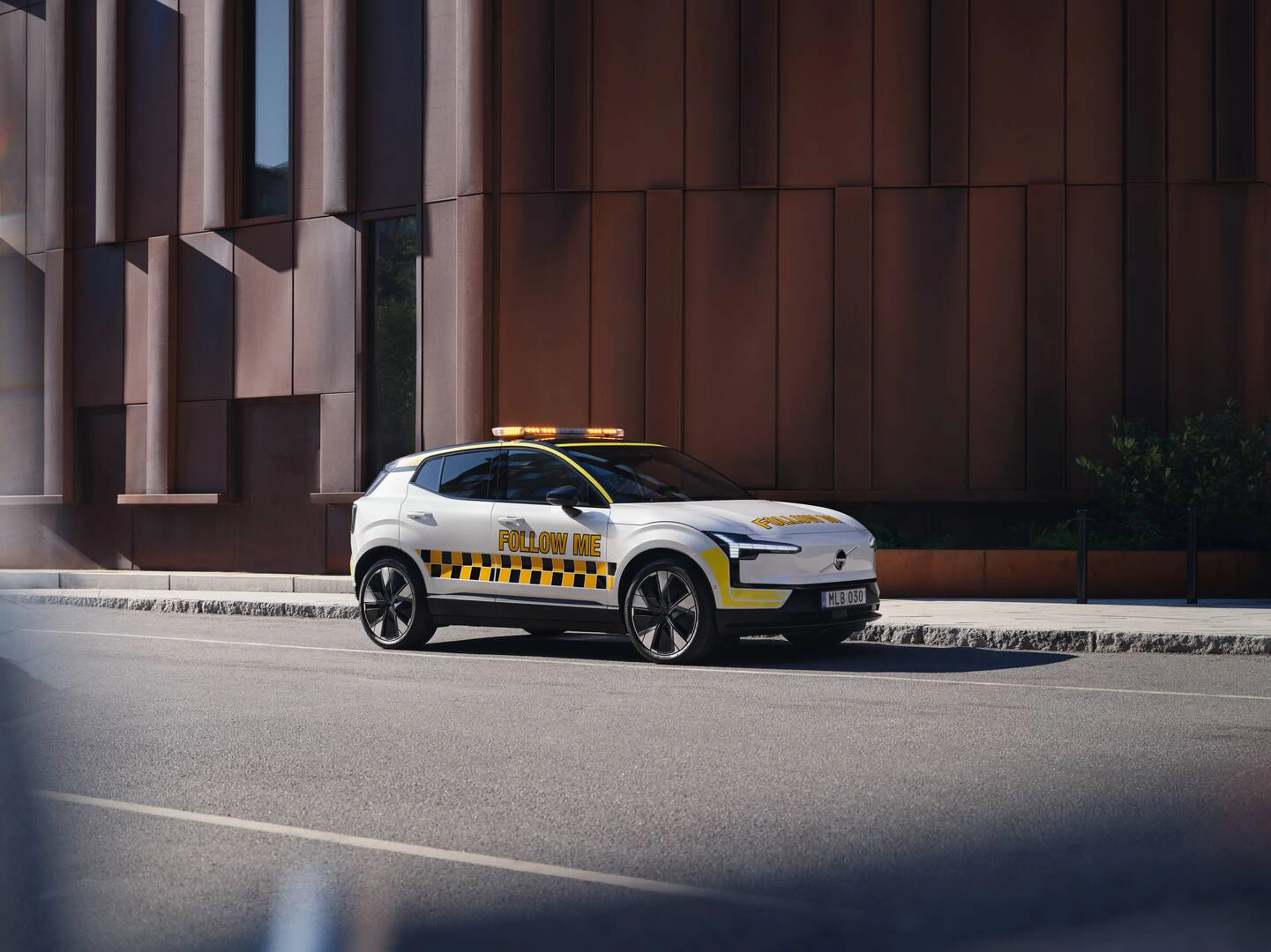 Автомобіль Volvo EX30 з картатими візерунками та помаранчевою світловою панеллю, припаркований біля будівлі аеропорту.