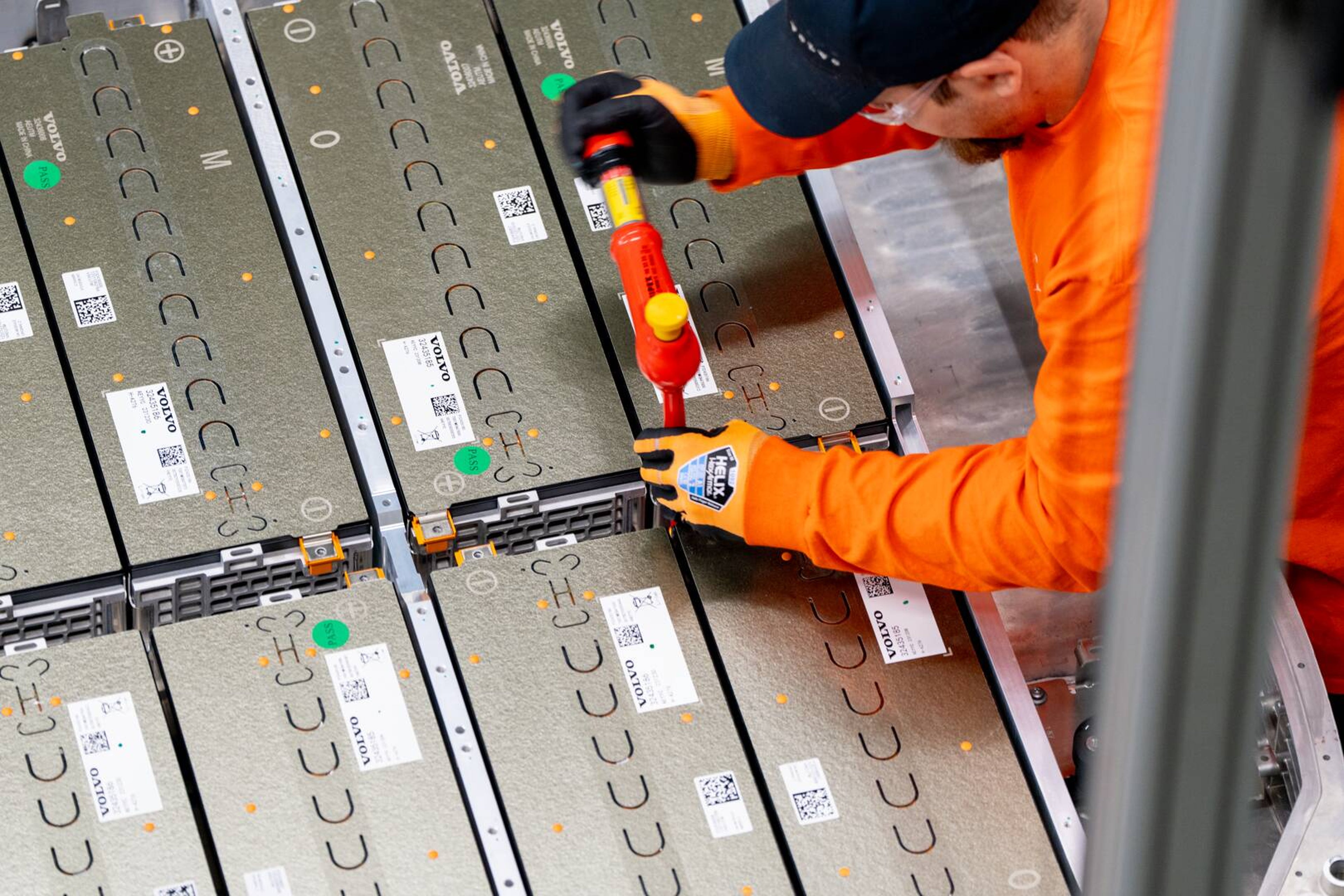 En arbejder iført orange sikkerhedsudstyr bruger et elværktøj til at samle eller fastgøre store batterimoduler, der er arrangeret i et gittermønster.
