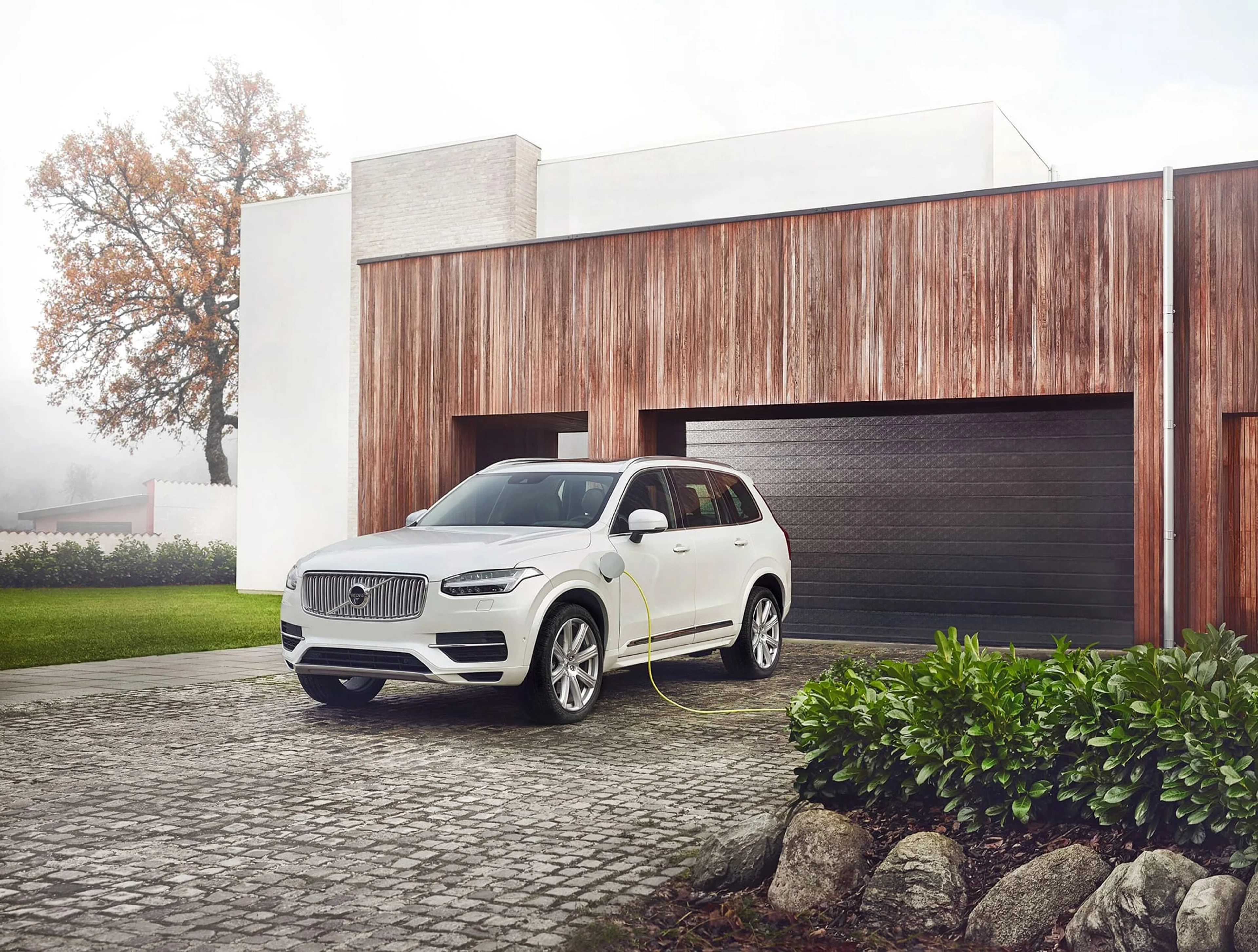 Une Volvo XC90 hybride rechargeable de couleur blanche, branchée pour la recharge, stationnée sur l'allée pavée d'une résidence moderne avec une façade en bois et un garage.