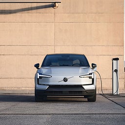 Vorderansicht eines Volvo Elektroautos, das an einem sonnigen Tag an eine öffentliche Ladestation auf einem Parkplatz angeschlossen ist.