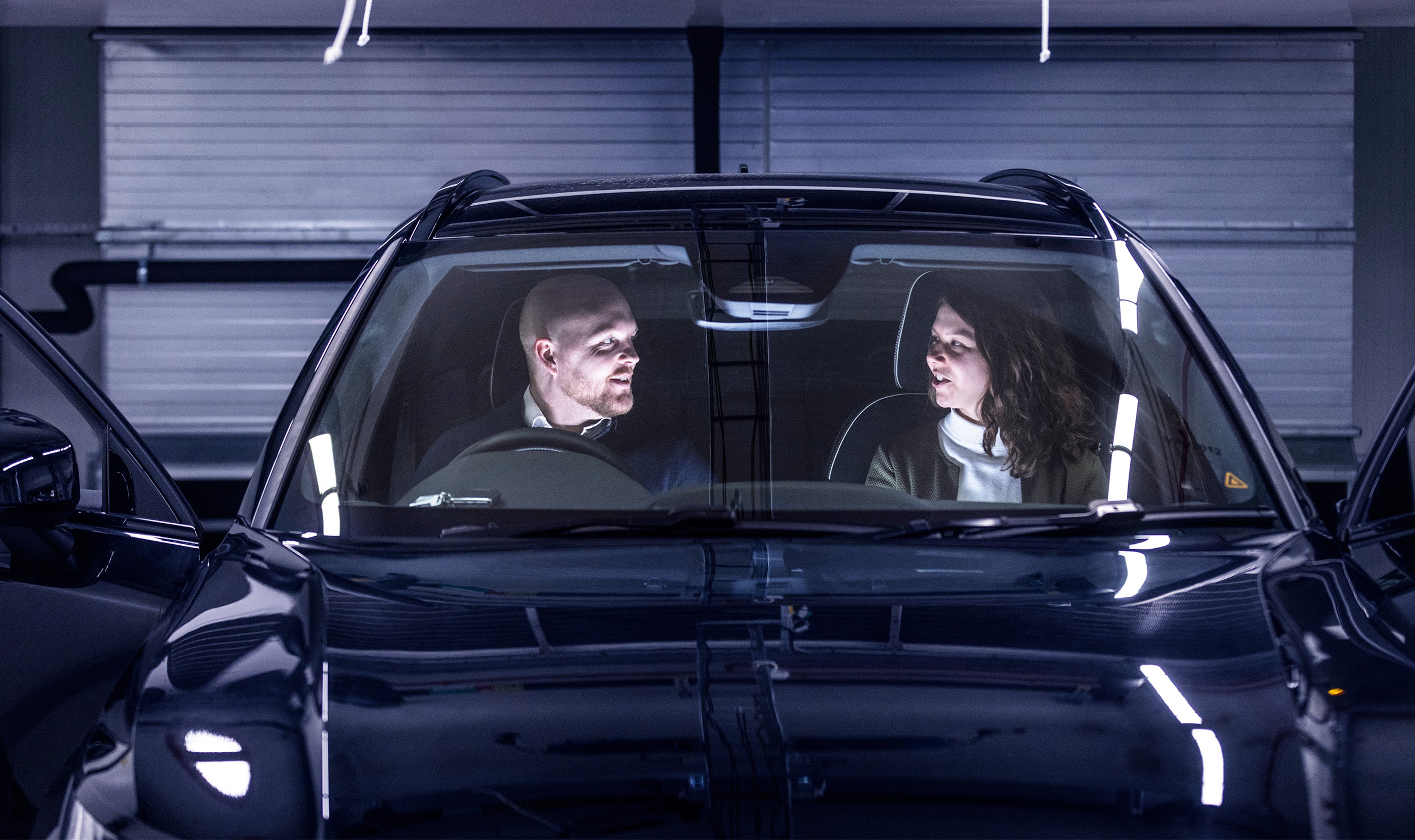 Vy framifrån av en svart Volvo XC90 parkerad inomhus med båda framdörrarna öppna, och visar två personer som sitter i fordonet. Strålkastare reflekteras på bilens vindruta och motorhuv.