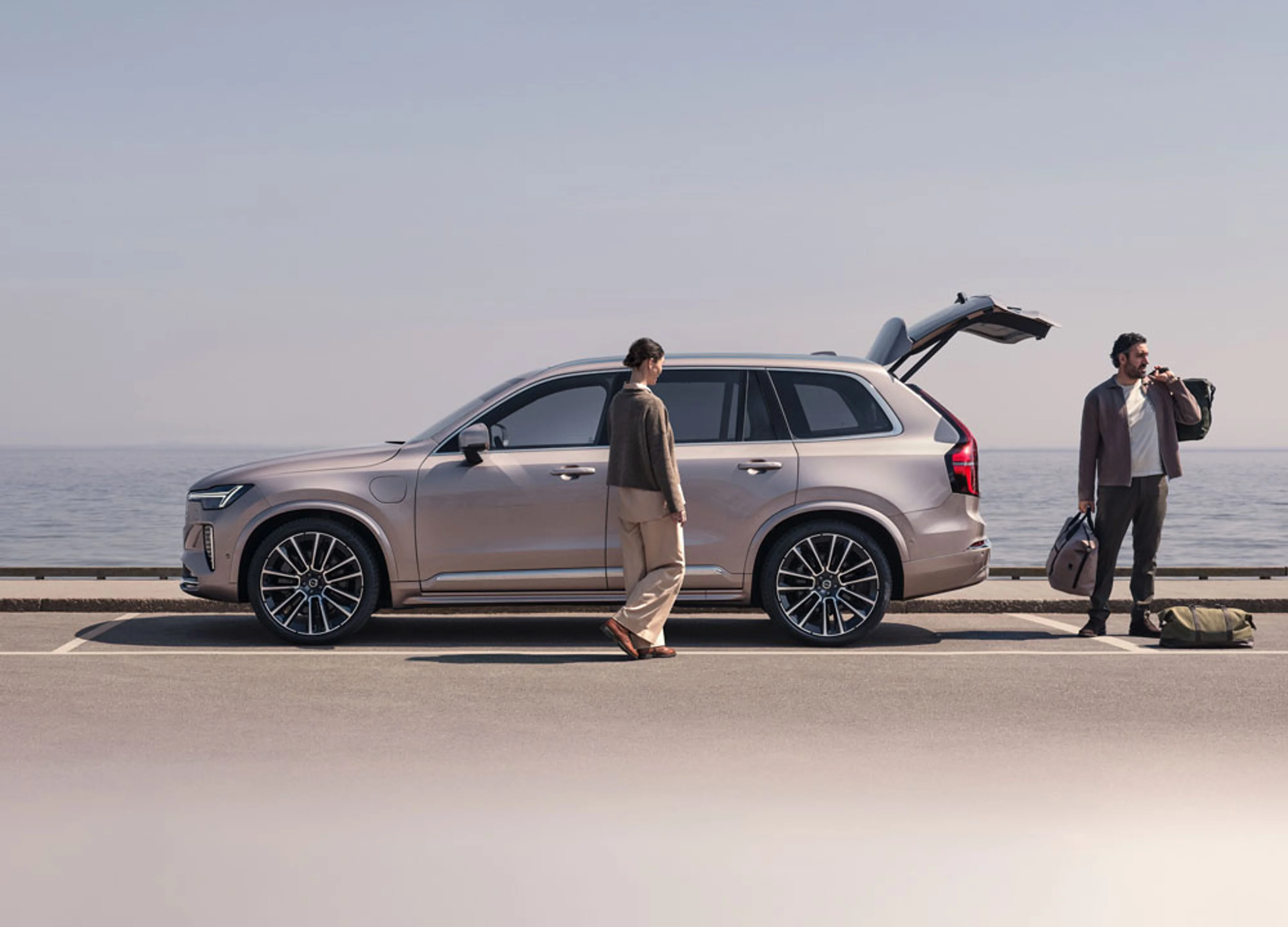 Deux personnes près d’un SUV Volvo beige au coffre ouvert, stationné sur une route côtière face à la mer.