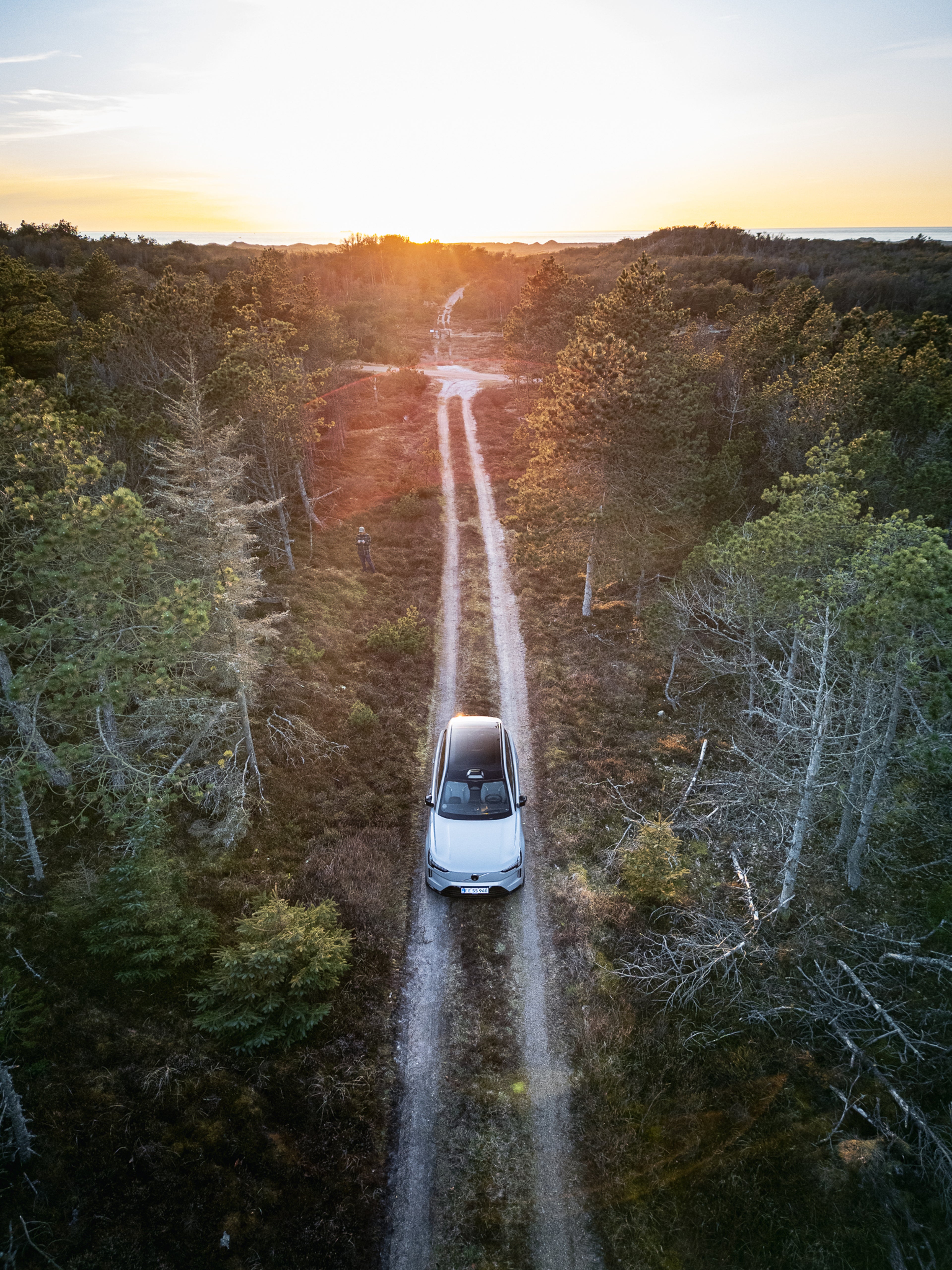 Luftfoto af en hvid Volvo bil, der kører på en smal grusvej gennem en skov ved solnedgang, omgivet af grønne træer. Scenen udstråler ro.