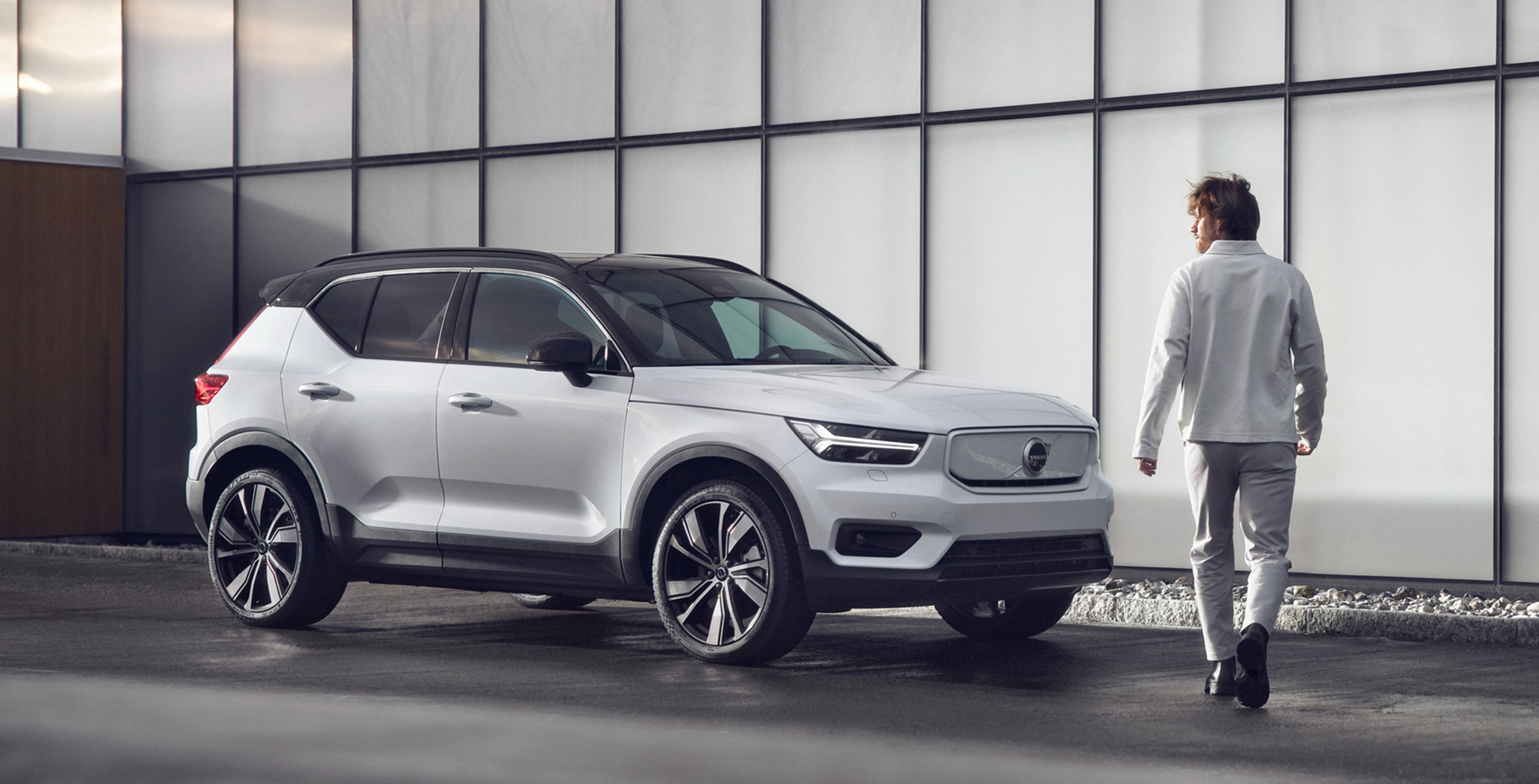 Vaaleisiin vaatteisiin pukeutunu mies kävelee kohti Crystal White pearl väristä Volvo XC40 Recharge P8 AWD autoa.