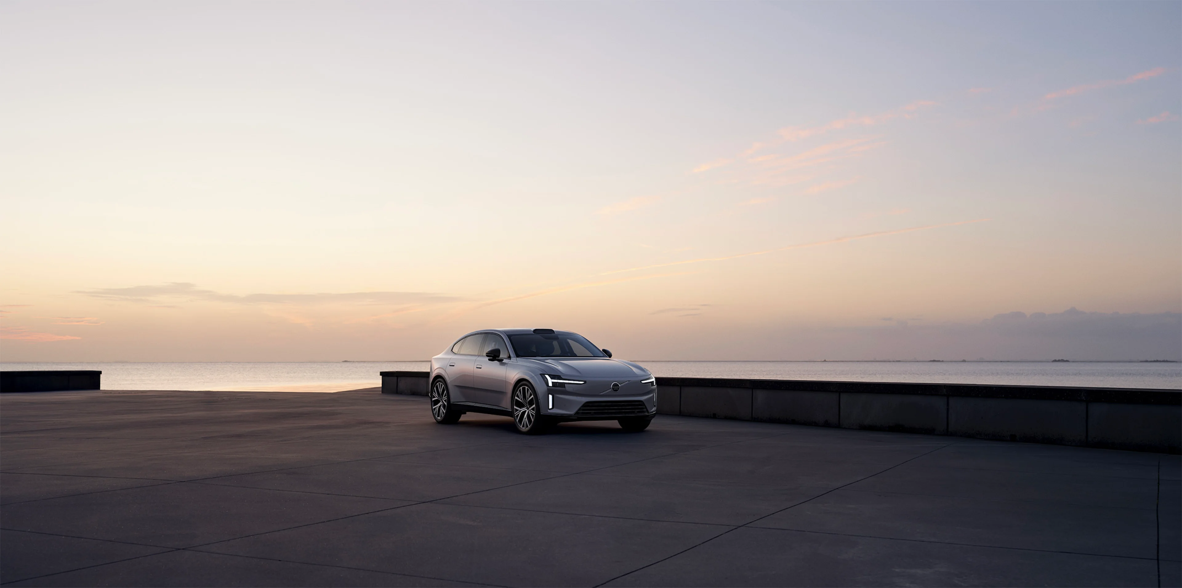Volvo ES90, naš sledeći potpuno električni automobil, parkiran na šetalištu pored vode u suton, prikazuje aerodinamički dizajn i LED farove.