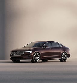Full exterior view of 2025 Volvo S90 Plug in Hybrid luxury sedan in burgundy parked in a modern city environment.
