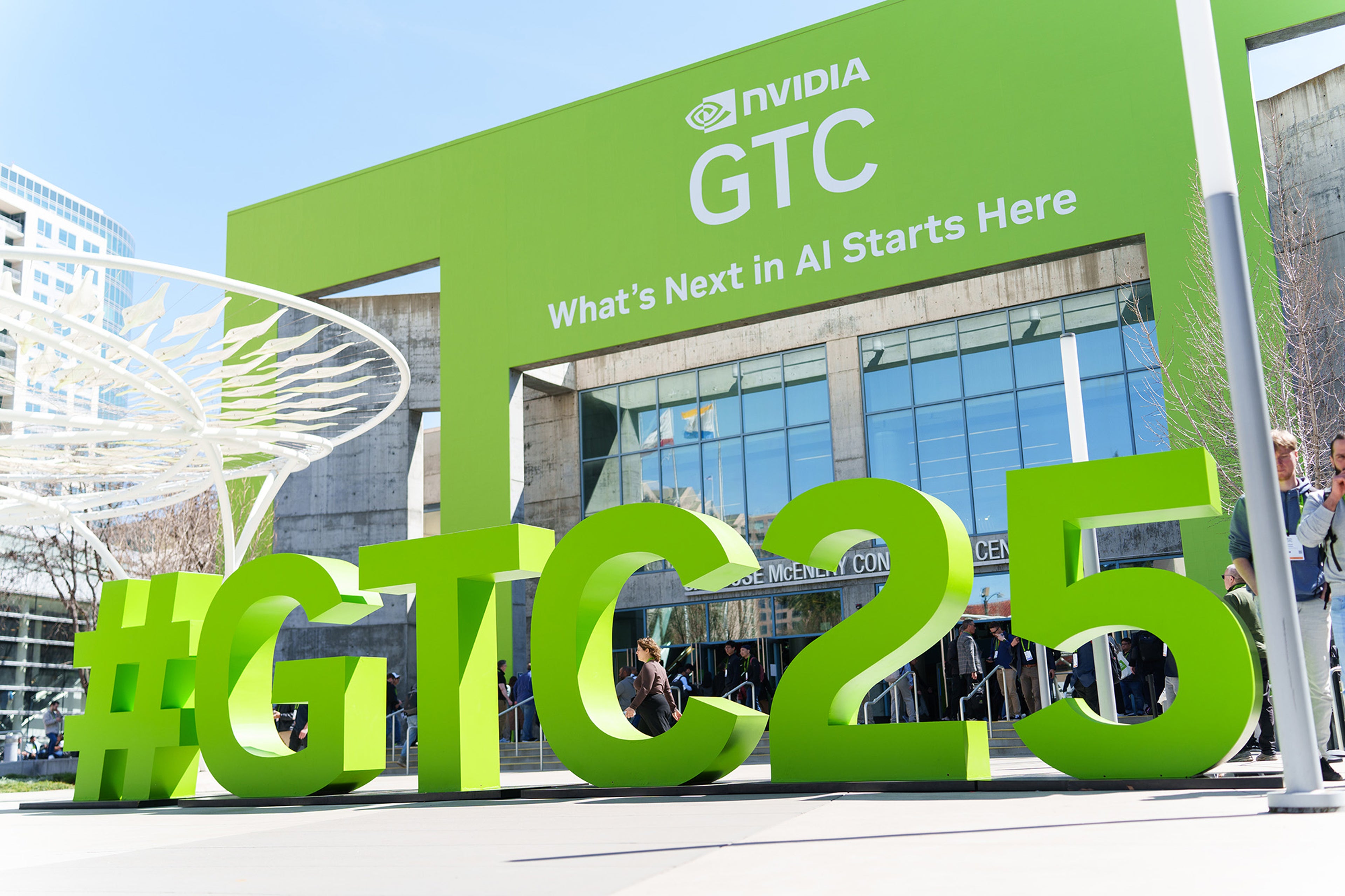 NVIDIA GTC 2025 event entrance with large green #GTC25 sign and 'What's Next in AI Starts Here' slogan at the San Jose McEnery Convention Center, featuring attendees arriving for the AI and technology conference.