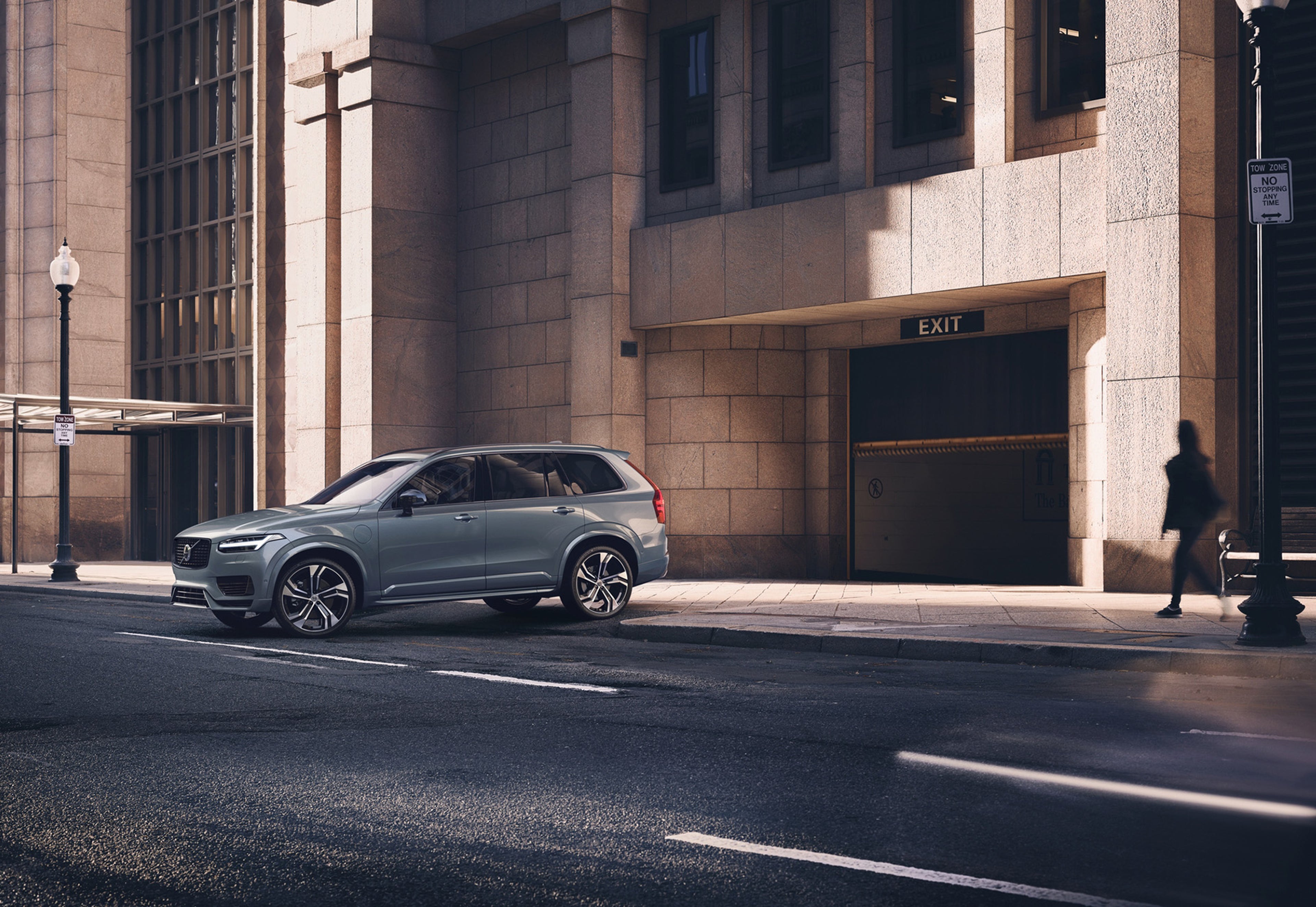 Un Volvo XC90 gris aparcado en una calle de la ciudad, frente a un gran edificio de piedra con una señal de salida sobre la entrada de un garaje. Una persona camina por la acera cercana.