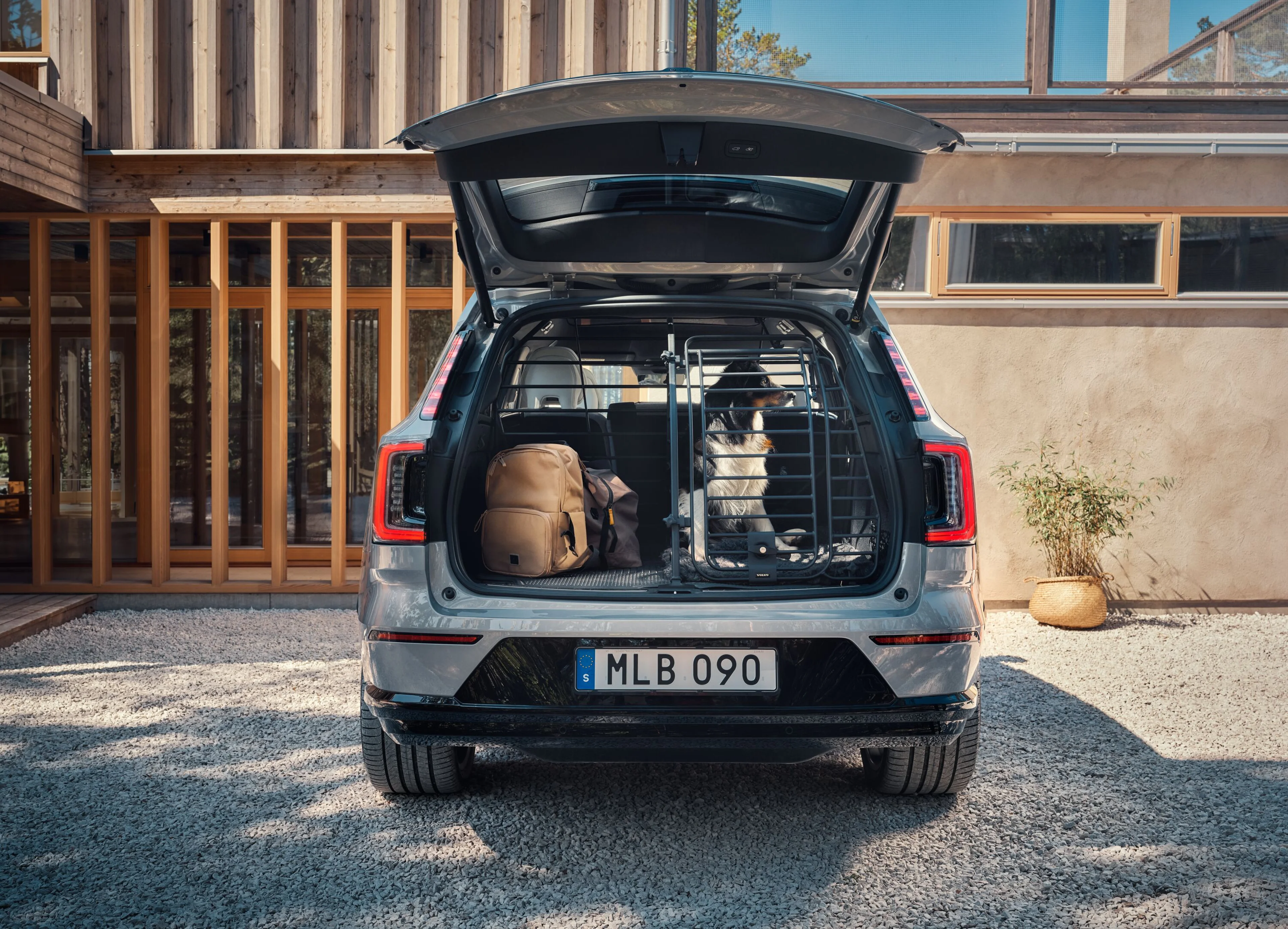 The open boot of a Volvo with a dog seated behind a dog gate, next to a backpack, in front of a house on a sunny day.