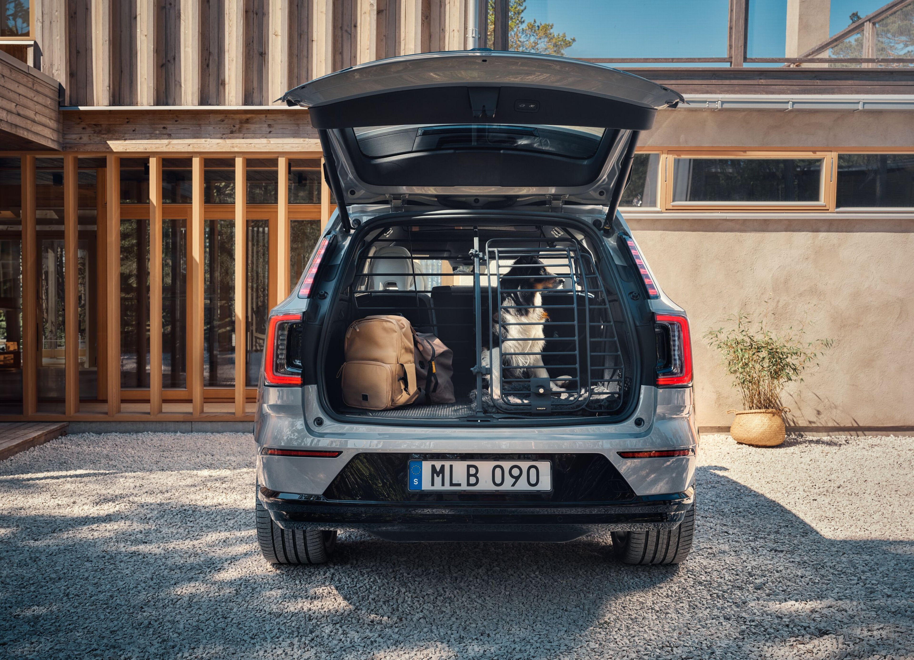 The open boot of a Volvo with a dog seated behind a dog gate, next to a backpack, in front of a house on a sunny day.