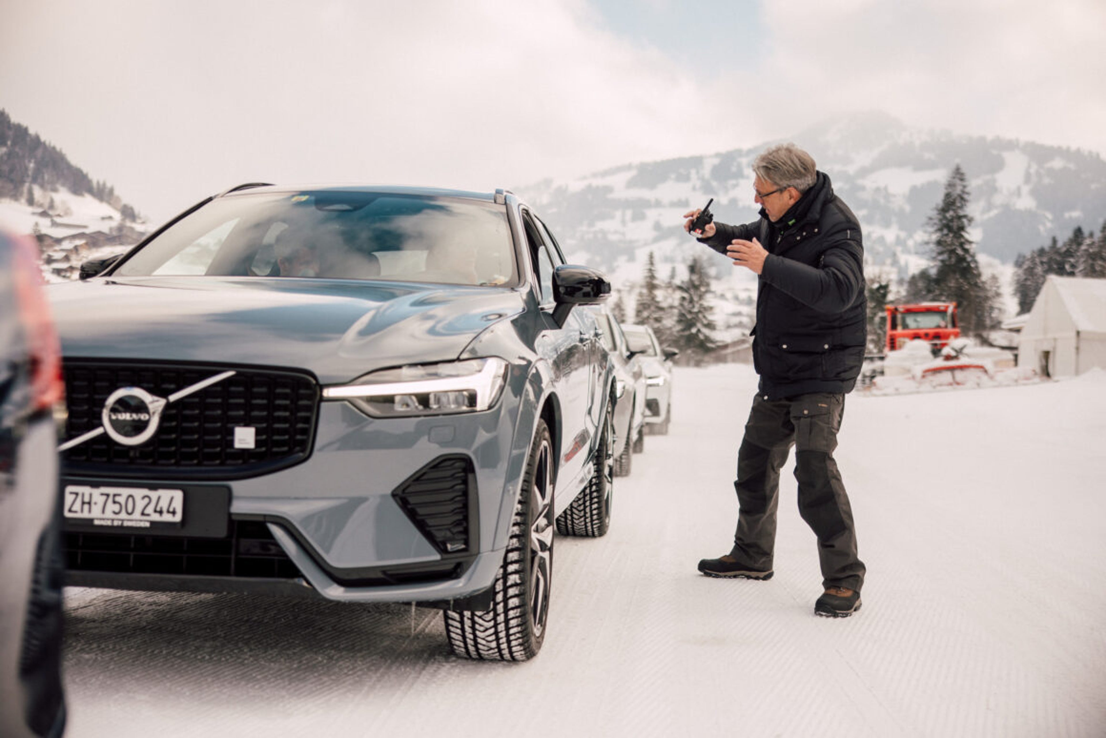 Ein mann steht mit einem Walkie-talkie vor einem grauen Volvo XC90 im Schnee.
