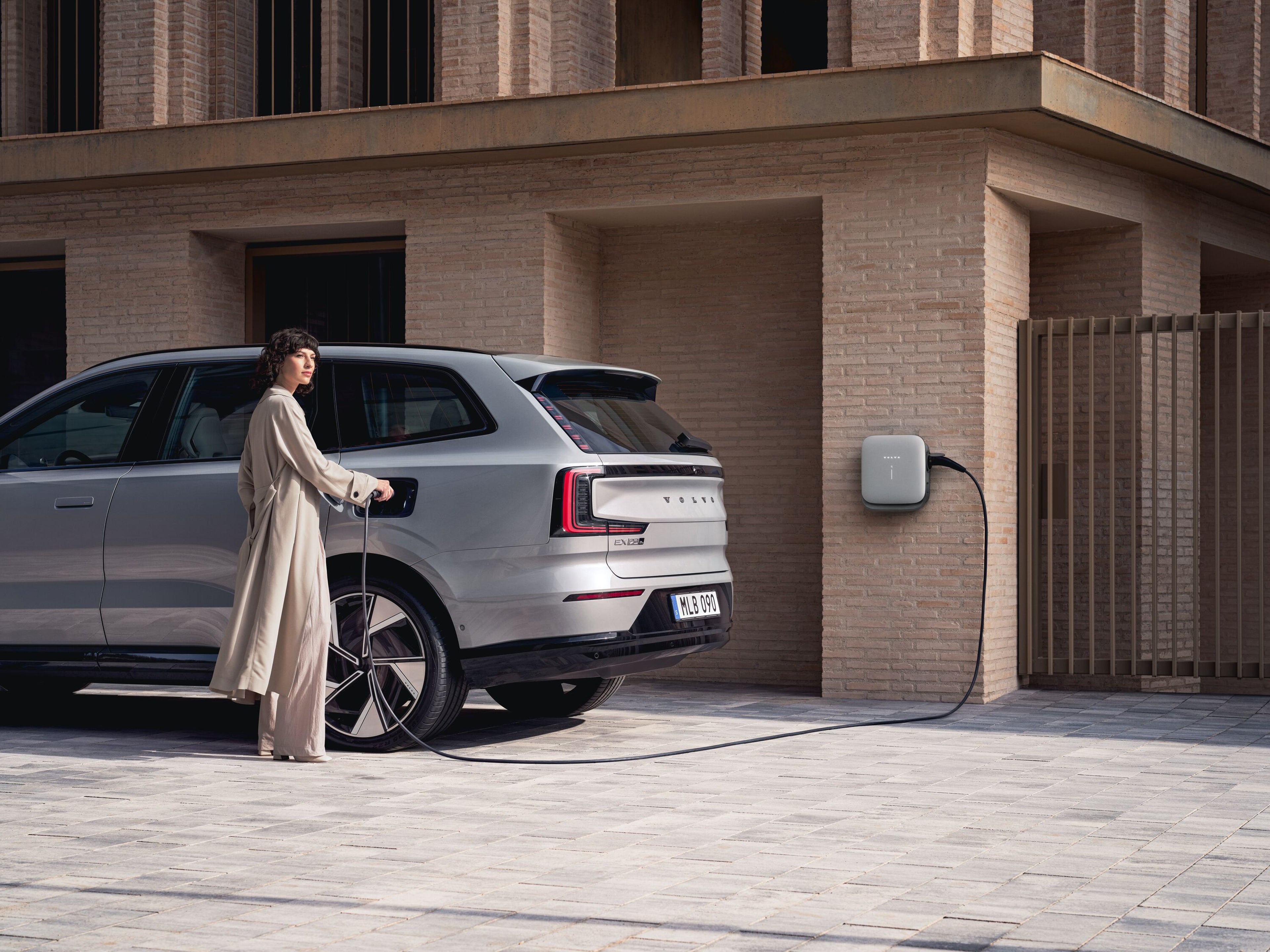 A female company car driver charging a Volvo EX90 at a wall-mounted station outside a brick building.