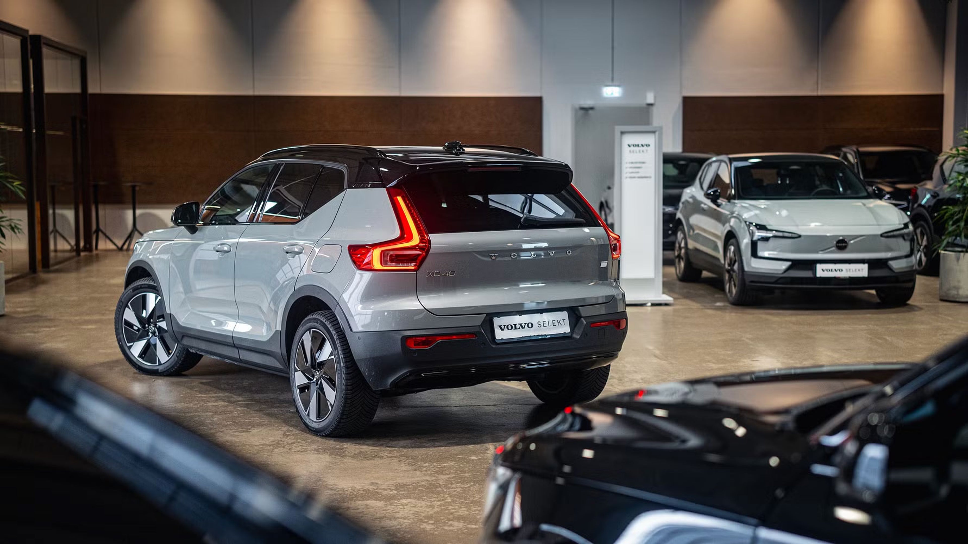 En grå SUV er udstillet i et showroom sammen med andre køretøjer. Synlig branding viser, at det er en Volvo. Belysningen er blød og fremhæver bilens elegante design.