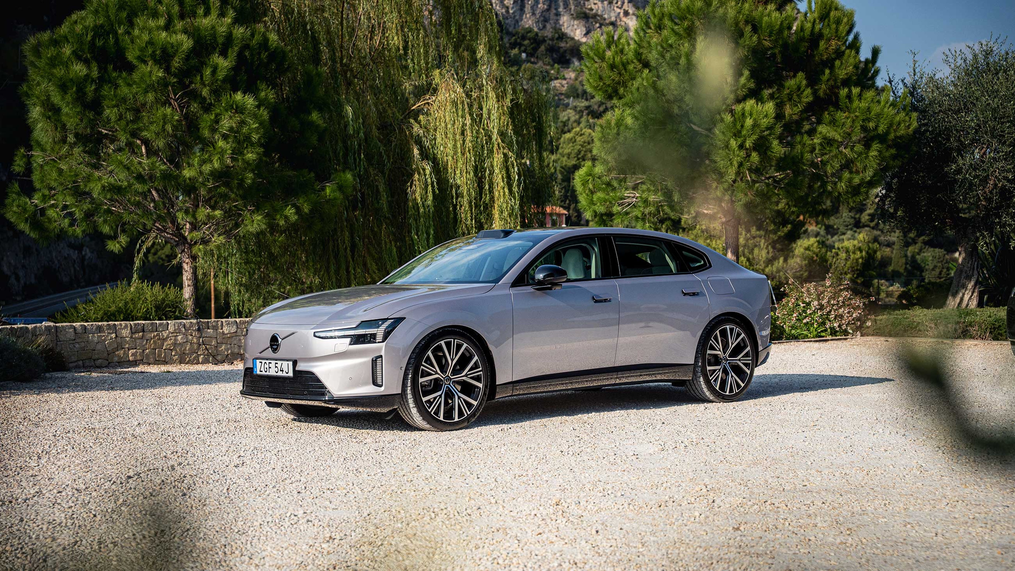 En sølvfarvet Volvo ES90 med slankt design og store hjul holder parkeret på grus, omgivet af frodige grønne træer. Scenen udstråler elegance og ro.