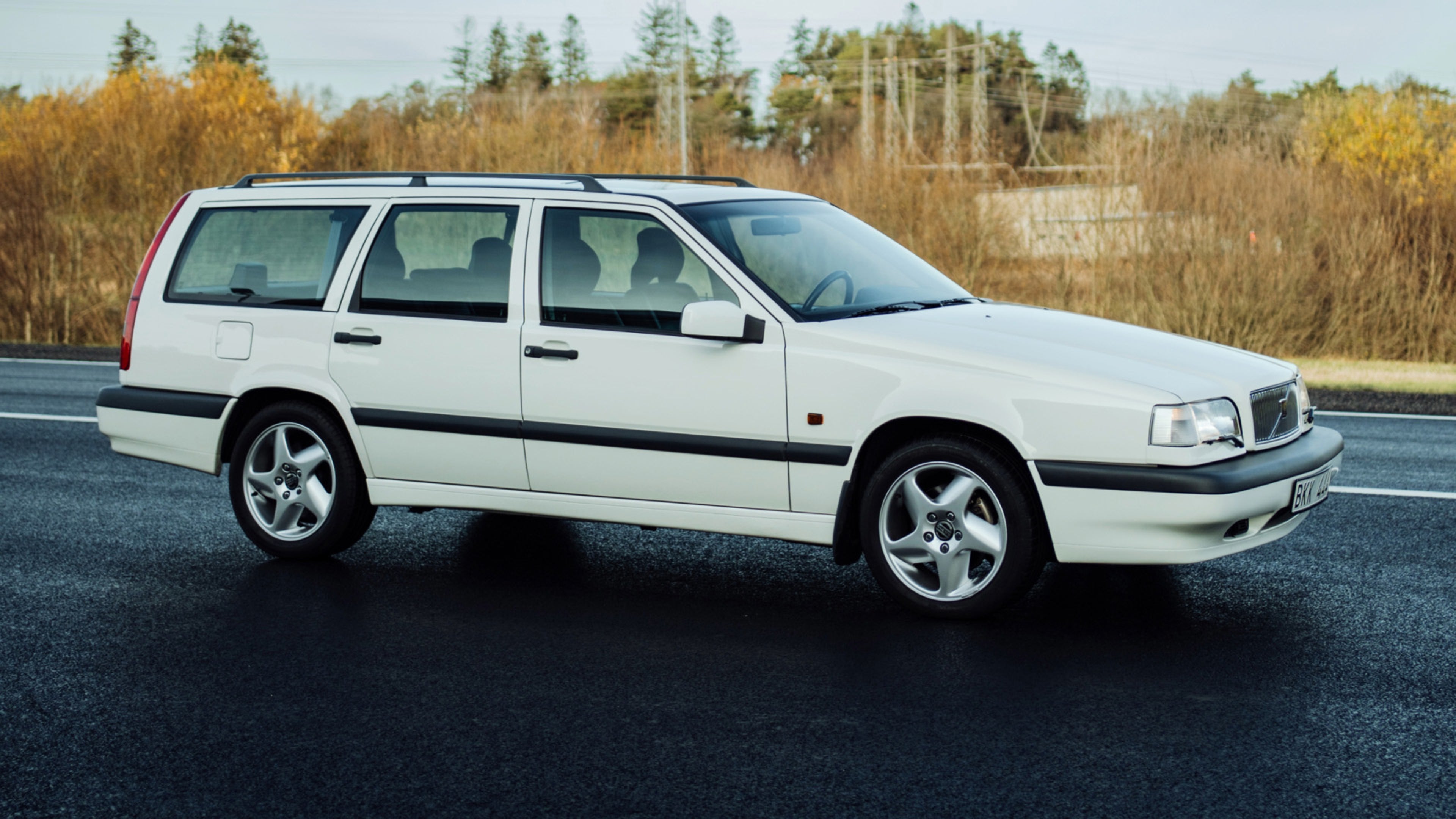 Vagon i bardhë Volvo 850 i parkuar në trotuar të lagësht me sfond të bardhë muri