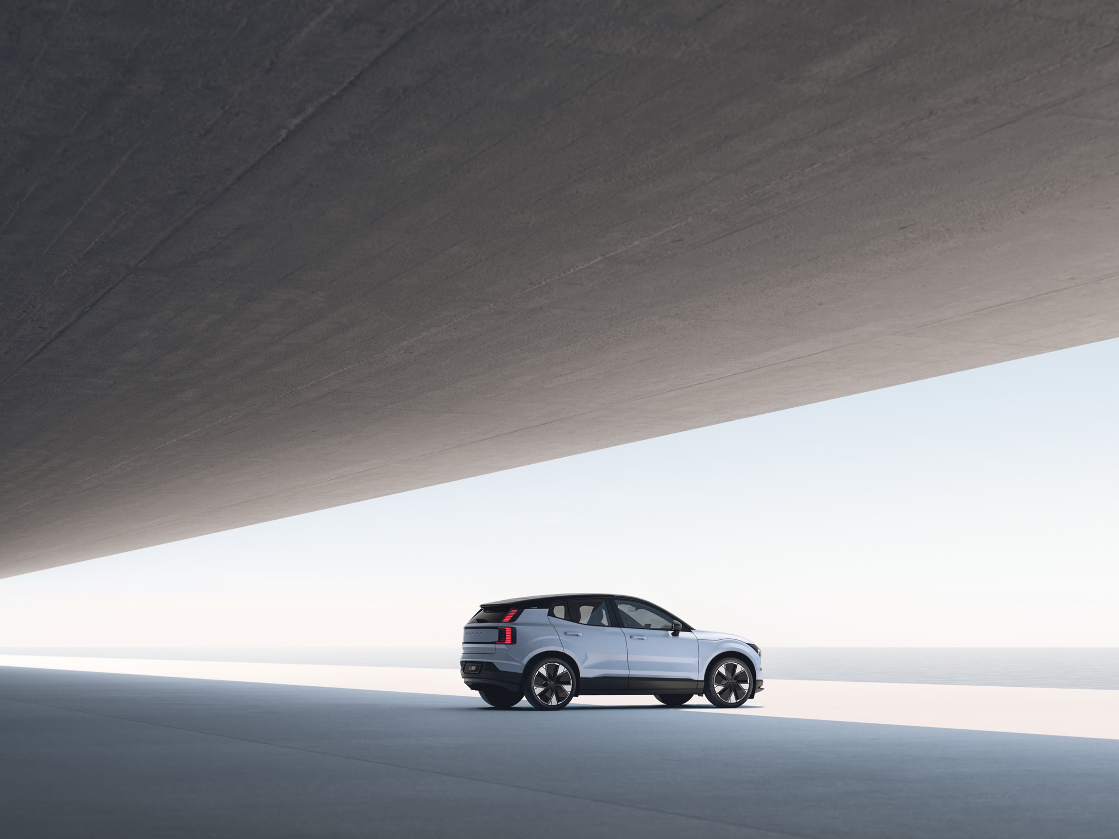 SUV EX30 argent garé sur une route côtière minimaliste, encadré par un énorme porte-à-faux en béton et offrant une vue dégagée sur l’océan.