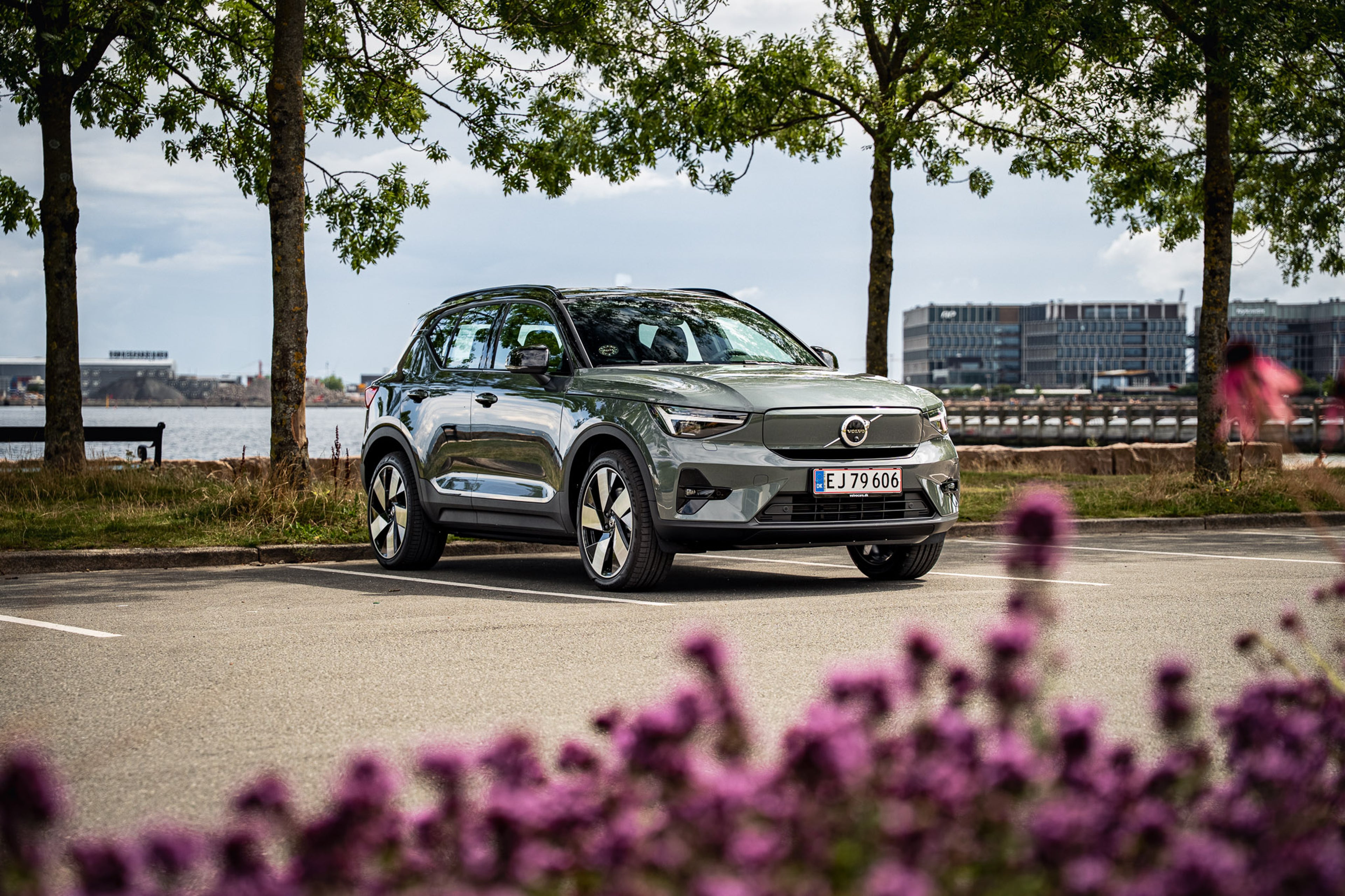 En elegant, grå Volvo bil holder parkeret ved en havnefront, omgivet af træer. I forgrunden er lilla blomster slørede, hvilket tilføjer et levende præg. Scenen udstråler ro.