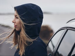 Egy kék kapucnis kabátot viselő nő profilképe, aki egy fehér Volvo mellett áll.