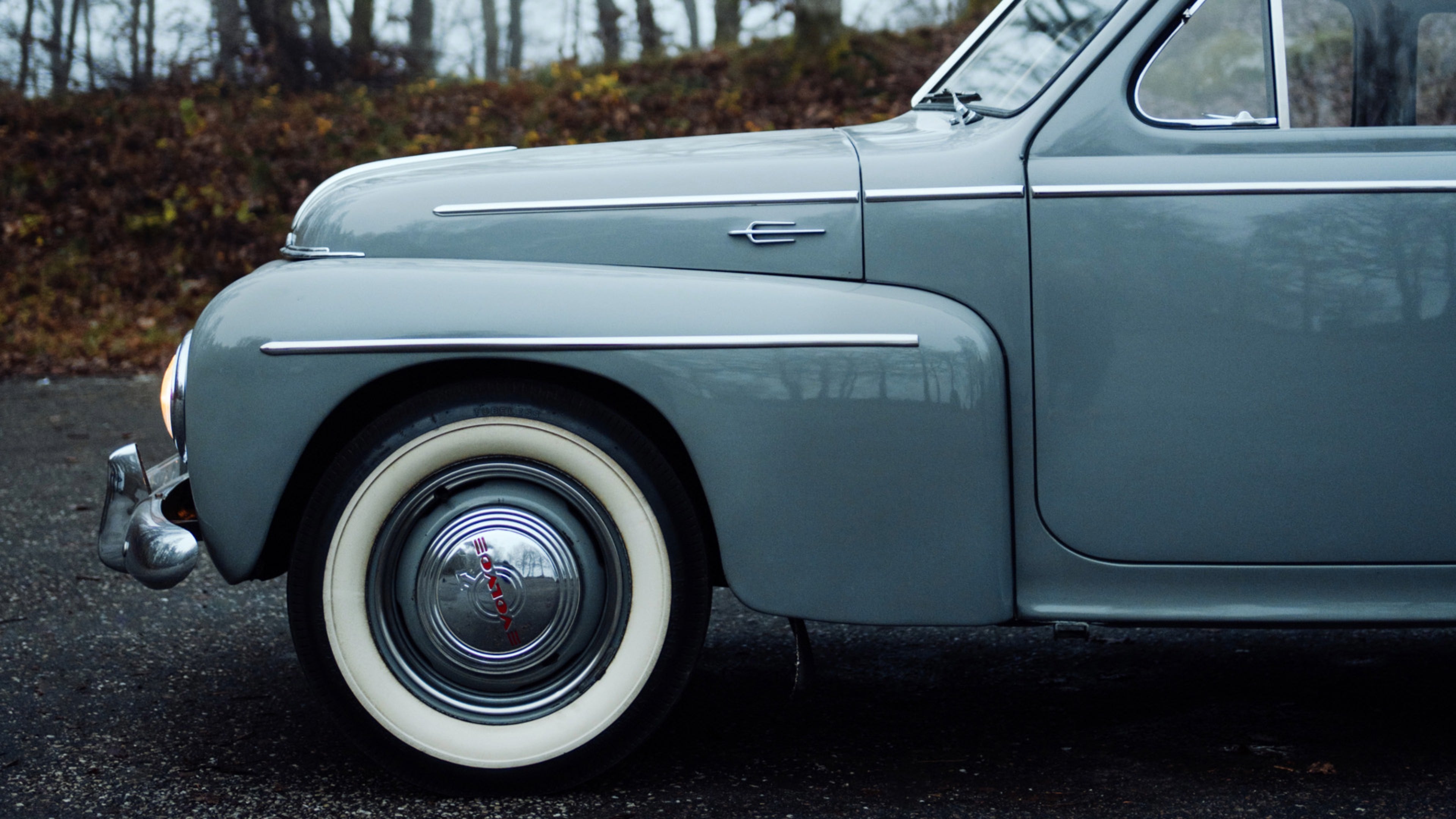 Страничен поглед на предната половина на класичен сив Volvo PV444 паркиран на влажен пат, на кој се гледа предниот браник, тркалото со раткапна на Volvo и дел од возачката врата со хромирани лајсни.