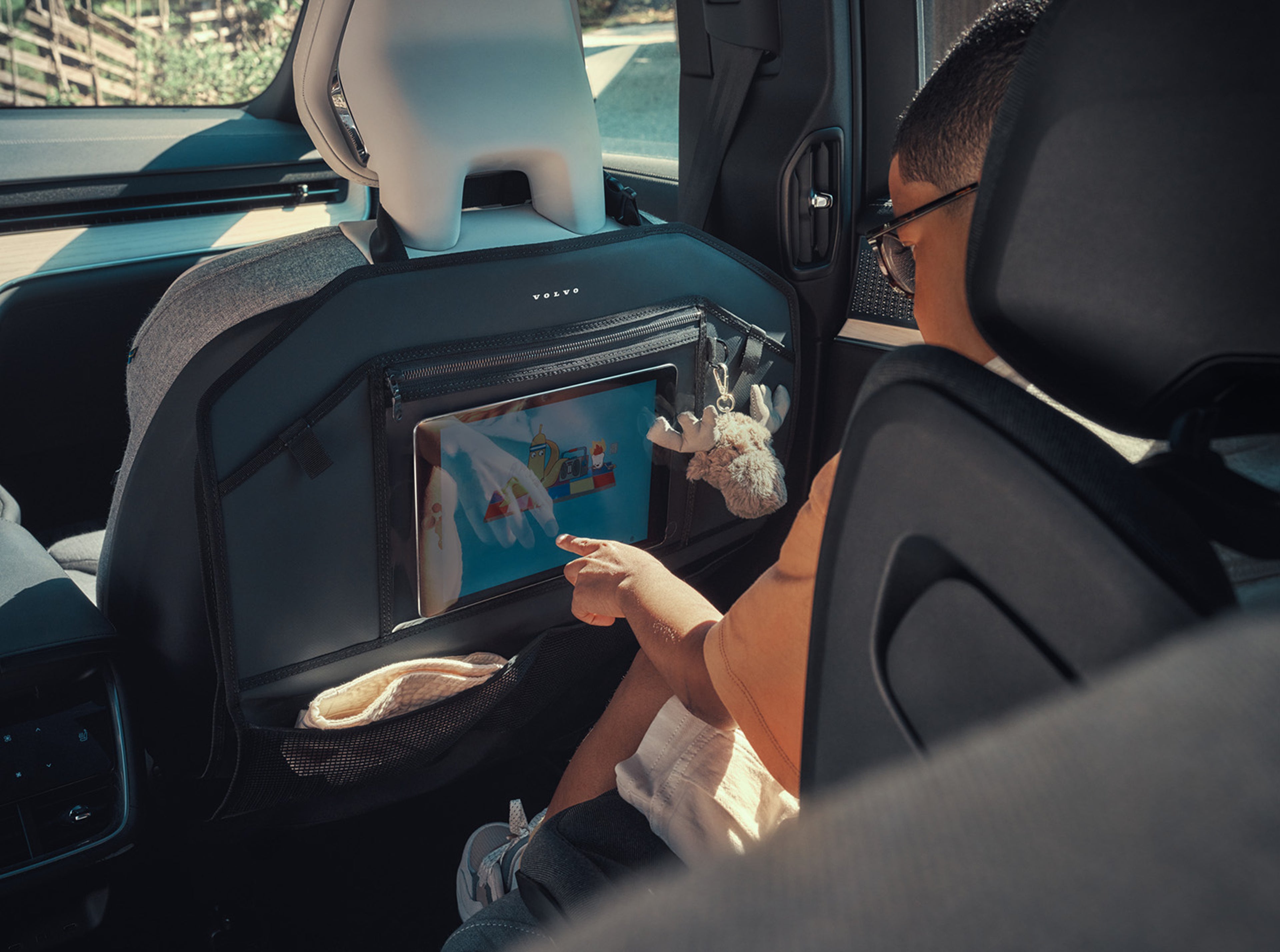 Een kind met een bril en een oranje shirt zit achterin een Volvo-voertuig en houdt zich bezig met een tablet die in een rugleuning is geplaatst. Een pluche sleutelhanger en opbergvakken zijn ook zichtbaar.
