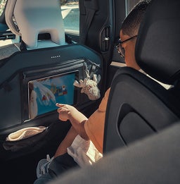 Een kind met een bril en een oranje shirt zit achterin een Volvo-voertuig en houdt zich bezig met een tablet die in een rugleuning is geplaatst. Een pluche sleutelhanger en opbergvakken zijn ook zichtbaar.