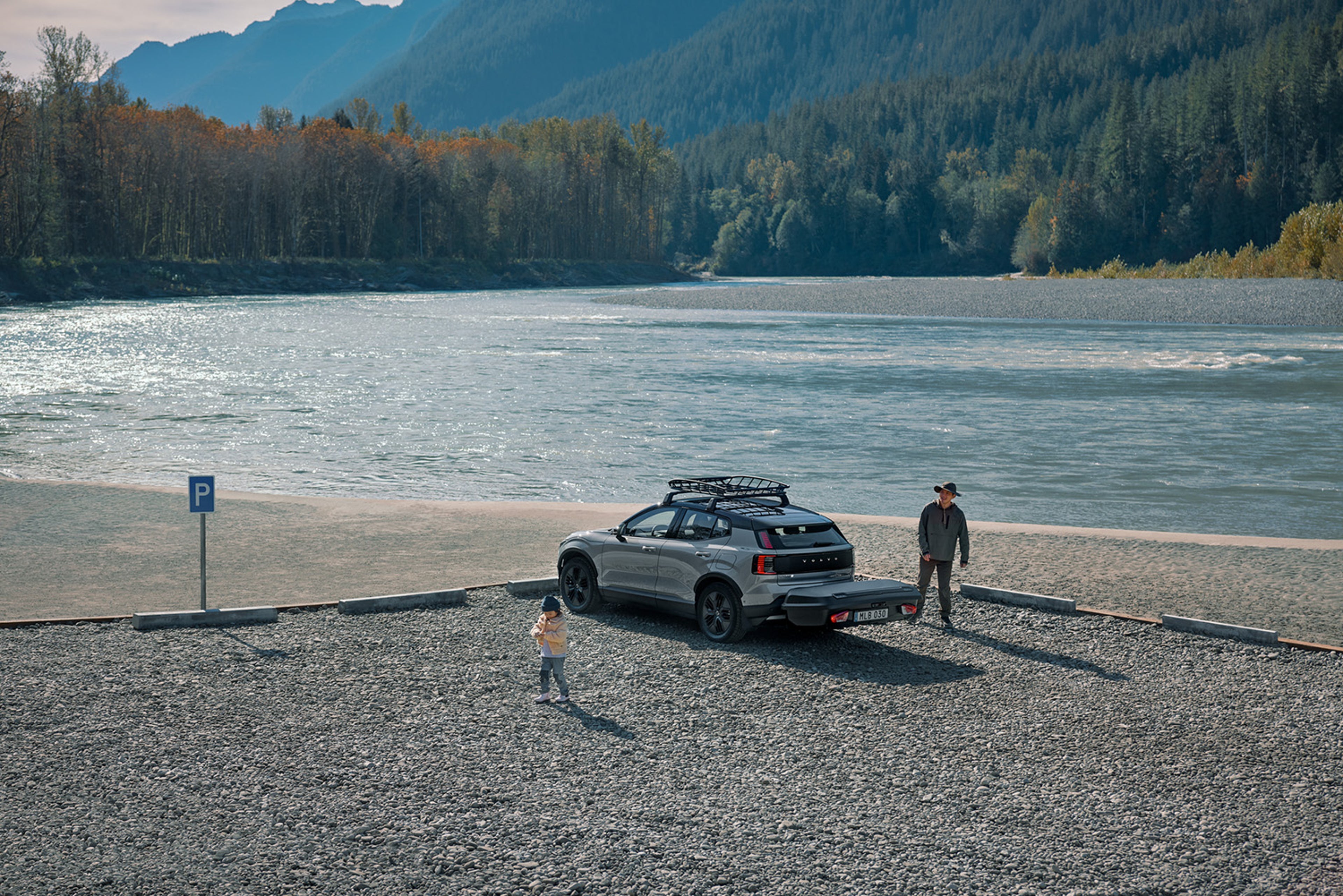 A Vapour Grey Volvo EX30CC with a roof rack is parked near a river, with a person wearing a hat standing beside it and a child playing in the foreground. Mountains and fall foliage are in the background.