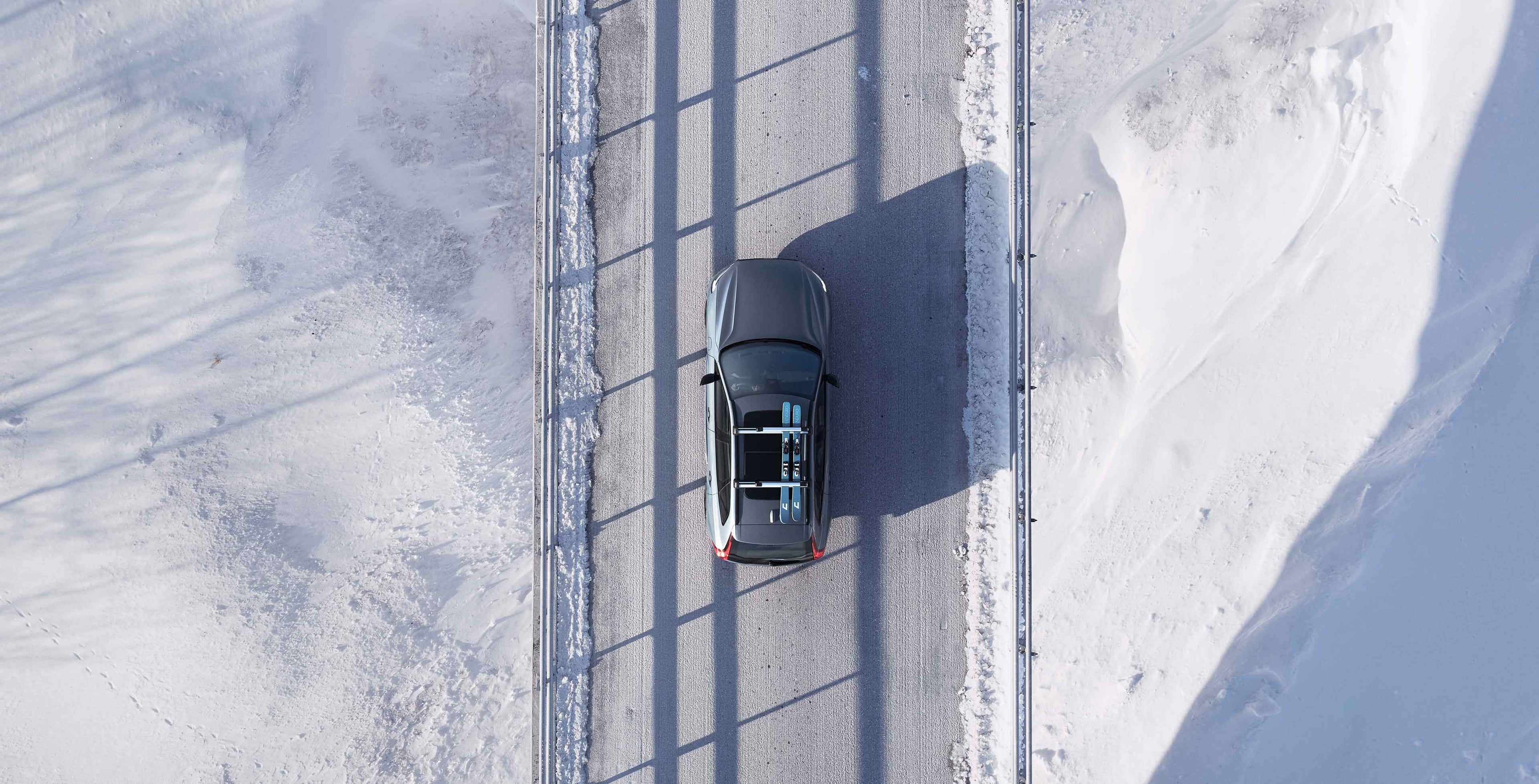 Ylhäältäpäin kuvattuna Volvo, joka ajaa talvisella tiellä pari suksia kiinnitettynä taakkatelineeseen.