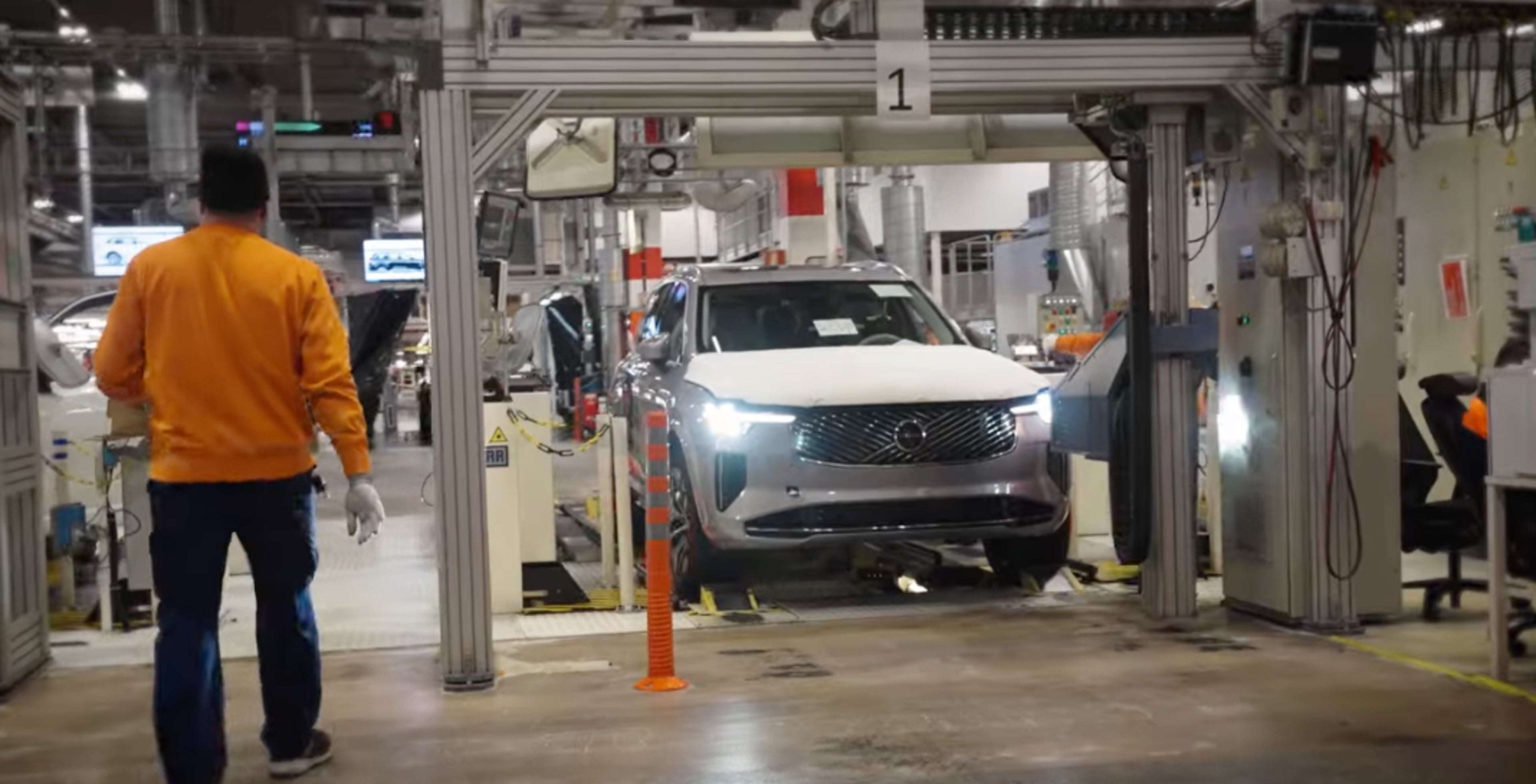 En arbejder i en orange jakke og handsker går hen imod en sølvfarvet Volvo bil på et samlebånd i en lyst oplyst fabrik, hvilket udstråler industri og præcision.