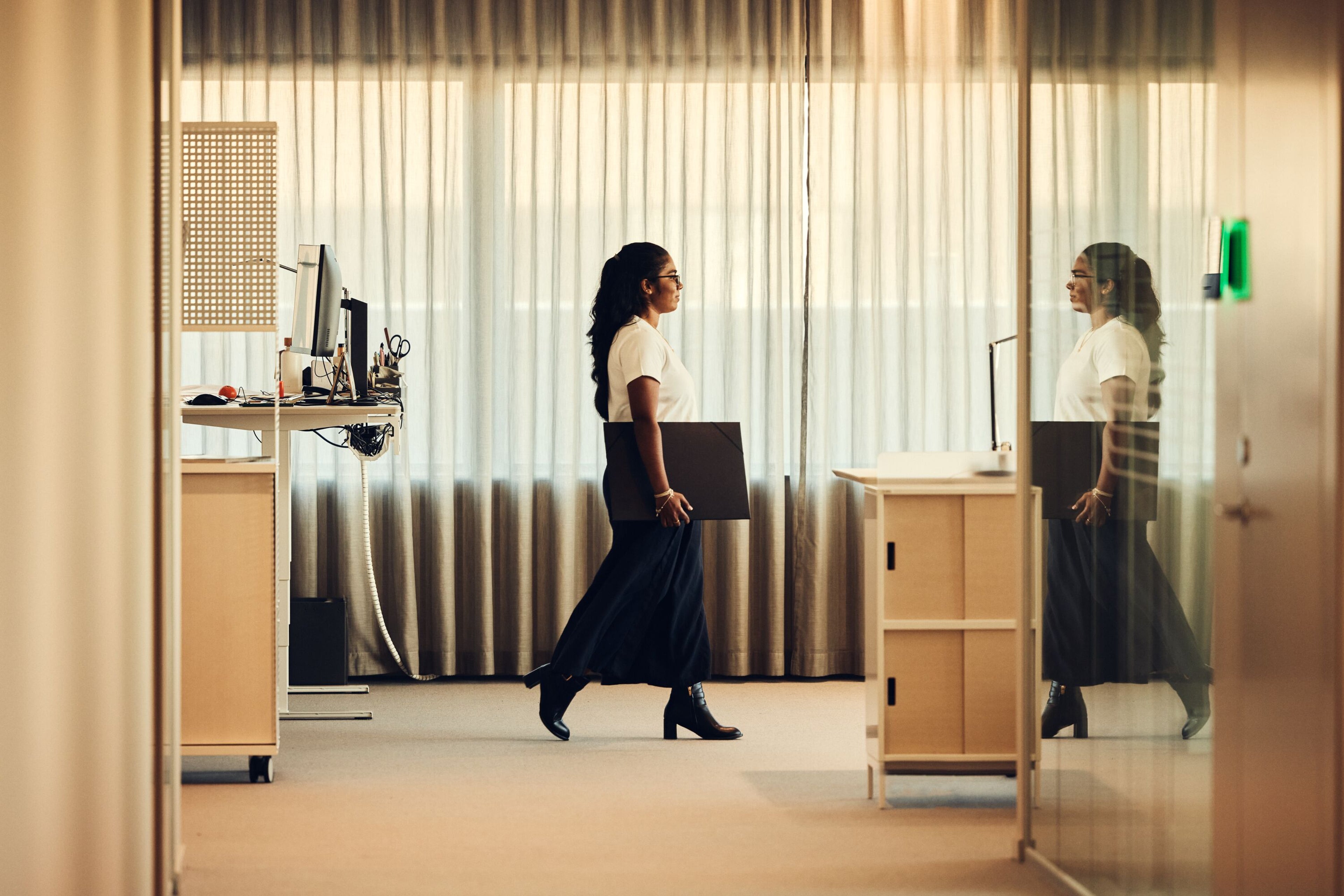A female fleet manager walks through an office holding a laptop beside a standing desk and curtained windows.