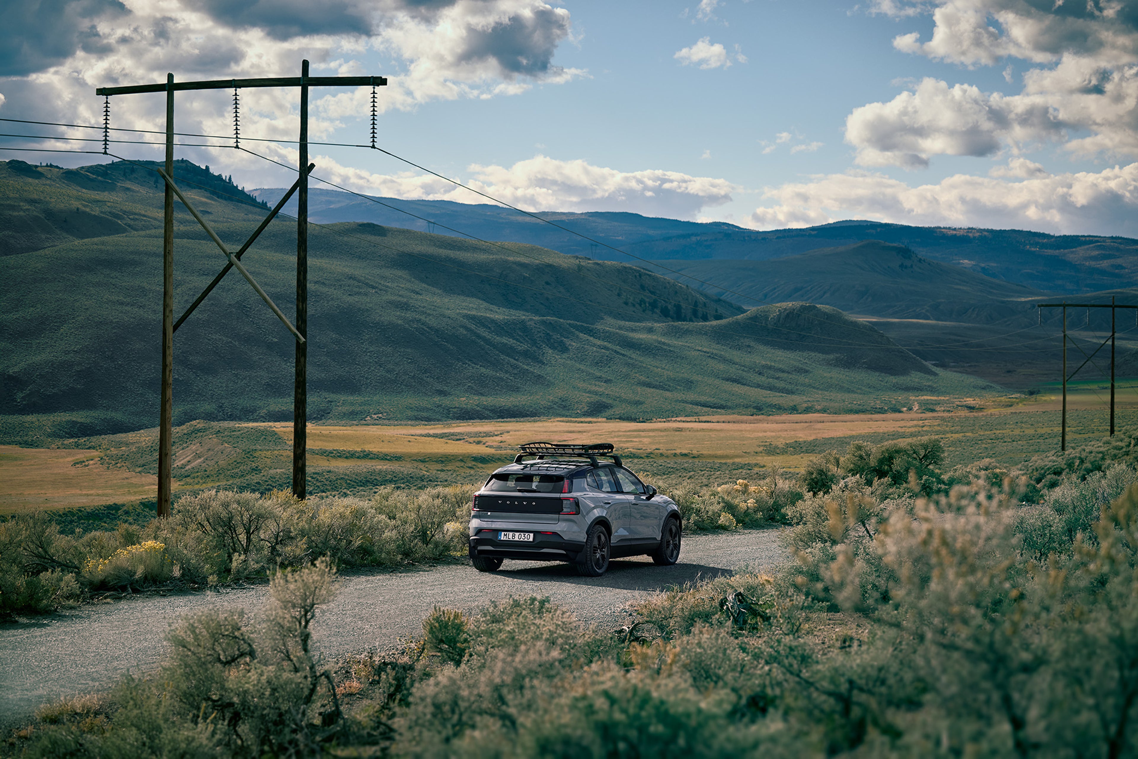 Un Volvo EX30 Cross Country Vapour Grey con barras de techo, circulando por una pintoresca carretera rural.