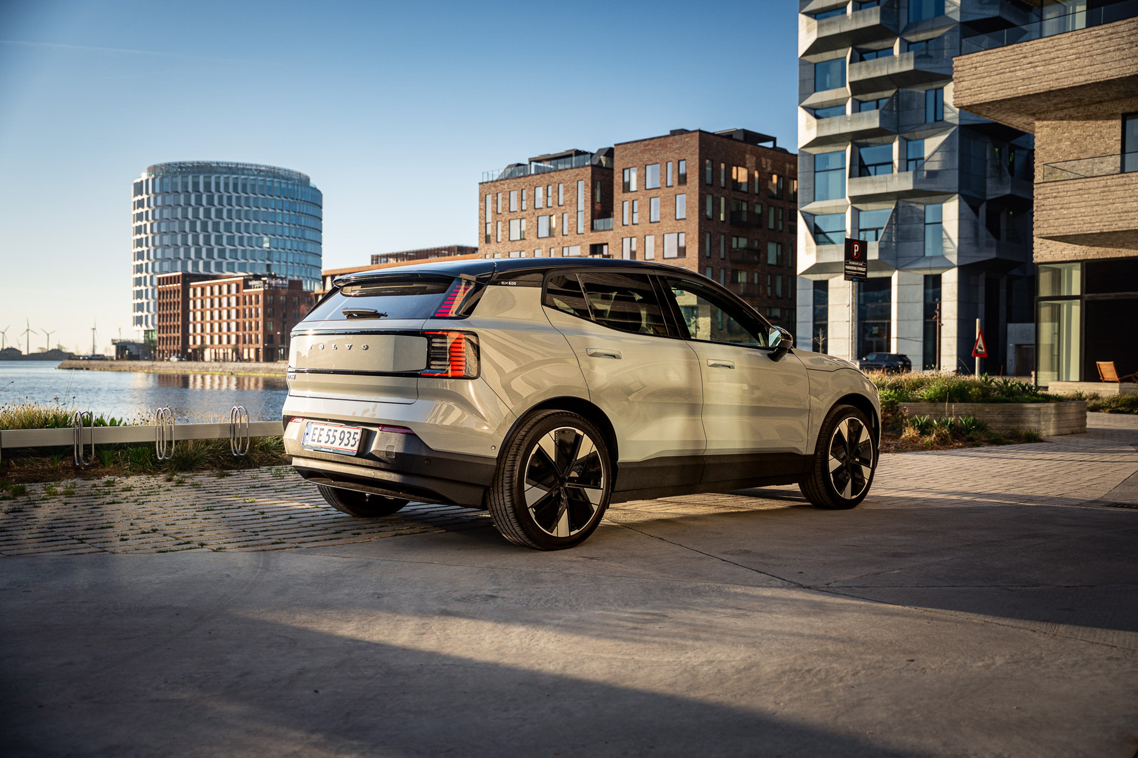 En elegant, moderne SUV med blank sølvfarvet lak holder parkeret nær bygninger ved vandet. Solen kaster et varmt skær, hvilket skaber en rolig, urban stemning.