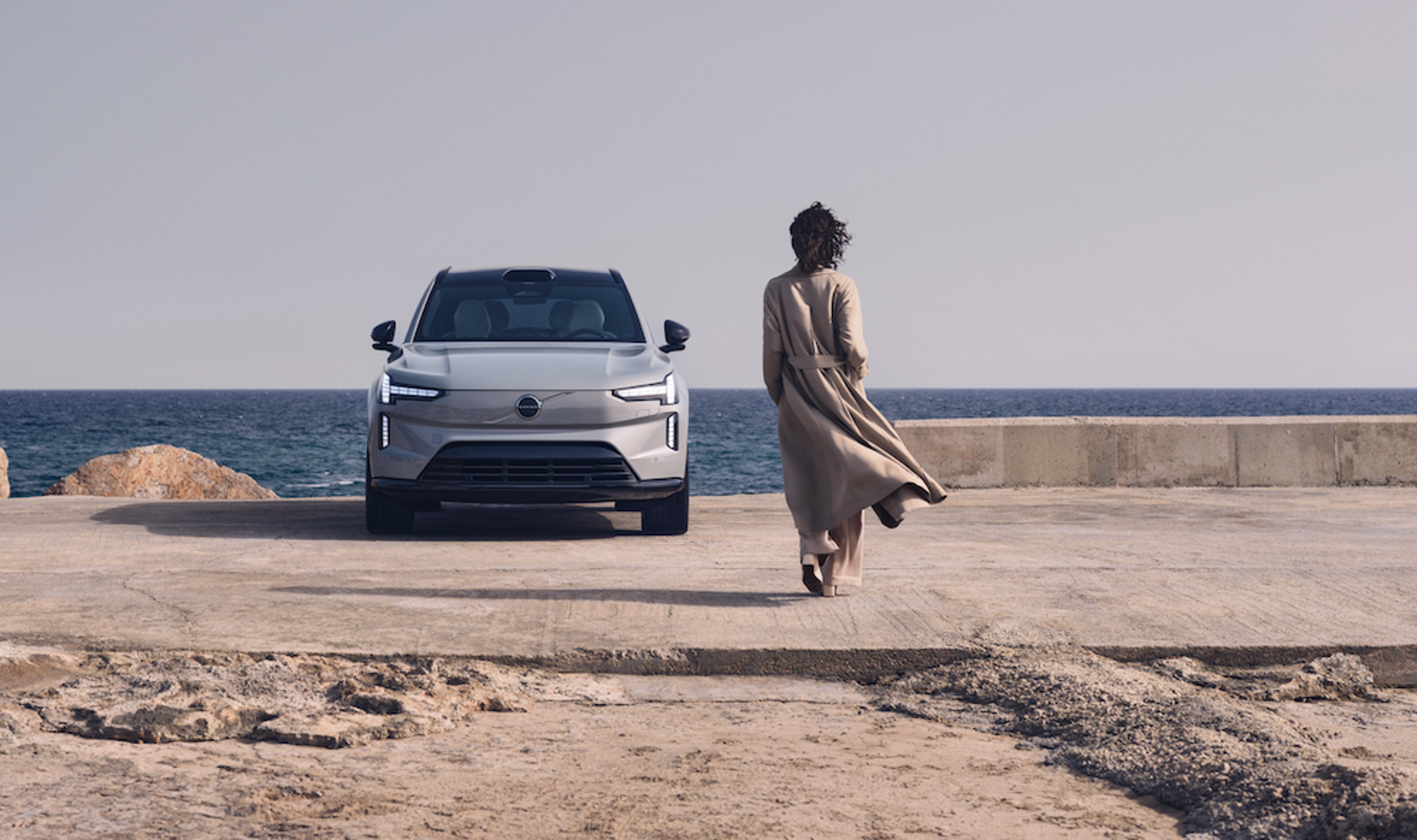 Ein grauer Volvo XC90 steht am Wasser. Eine Frau bewegt sich darauf zu.