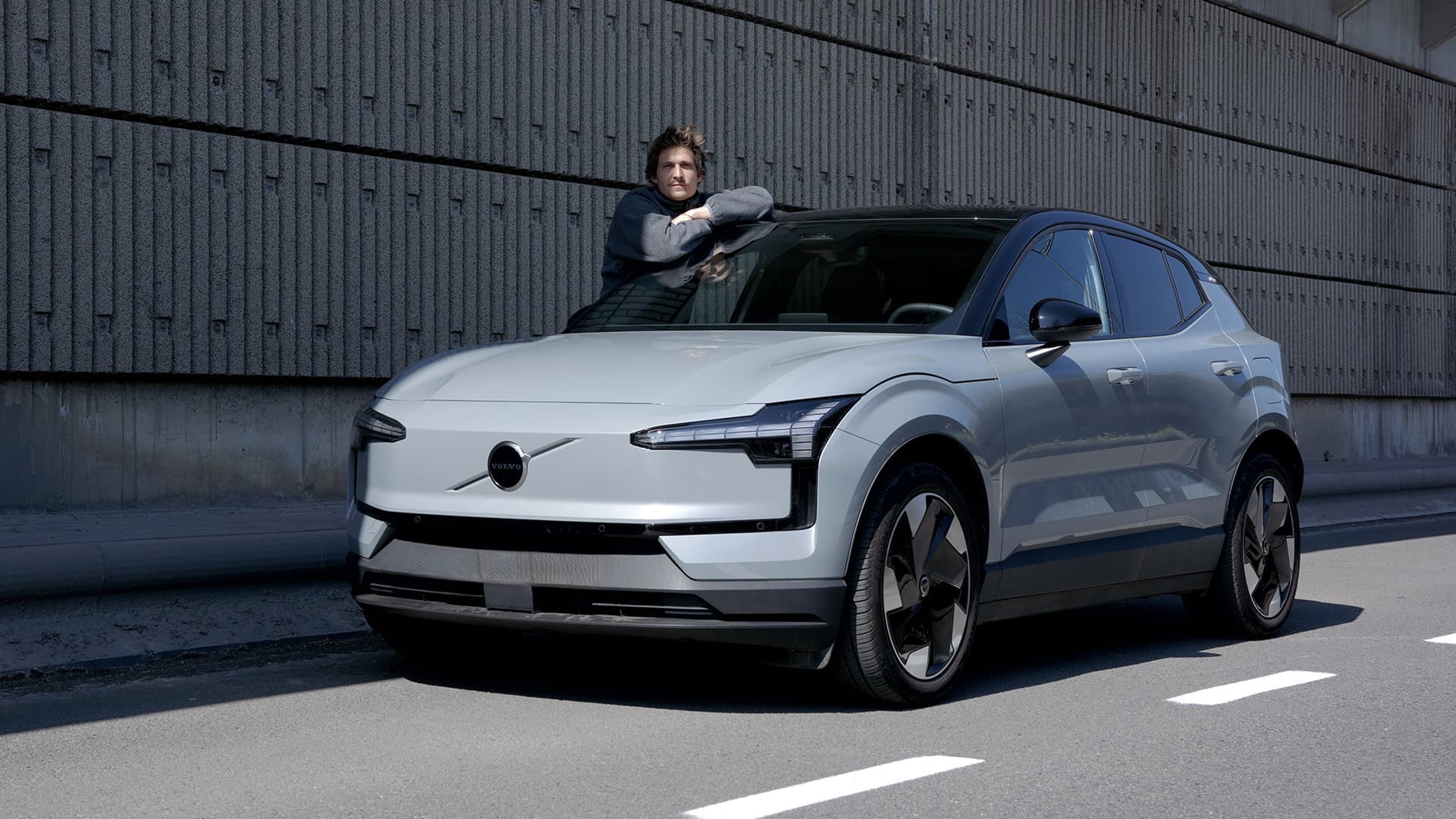 Jules Wittock est appuyé sur le capot d'une Volvo EX30 grise garée dans une rue avec un mur de béton gris en arrière-plan.