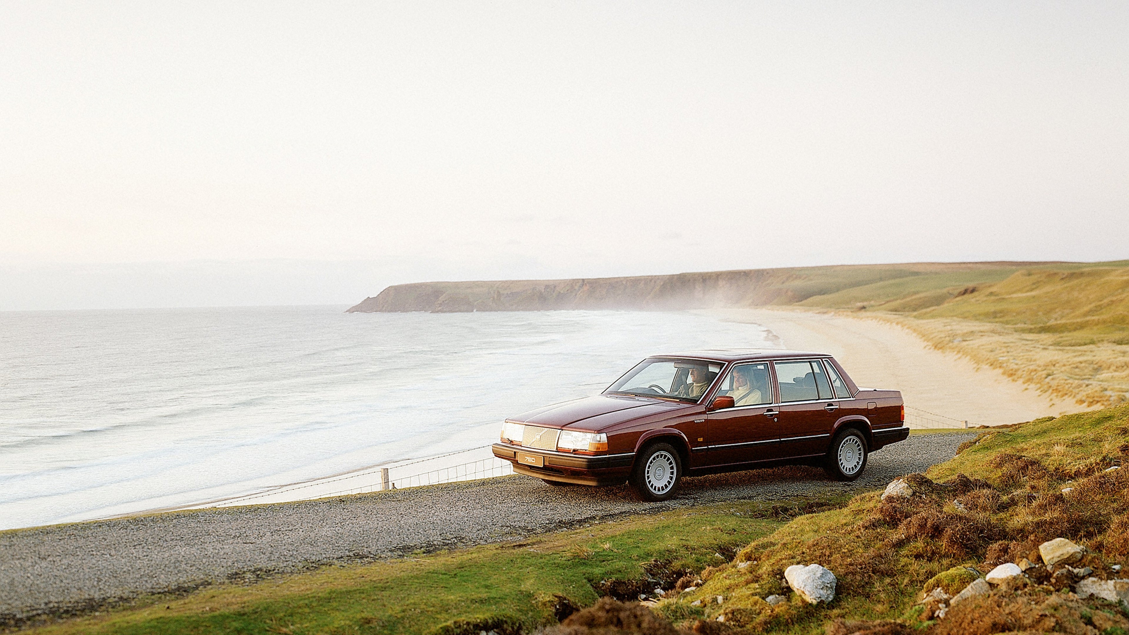 Volvo 760 GLE سيدان بنية مركونة على طريق ساحلي من الحصى مع أمواج المحيط والخط الساحلي الصخري والتضاريس العشبية والجرف البعيد تحت سماء صافية