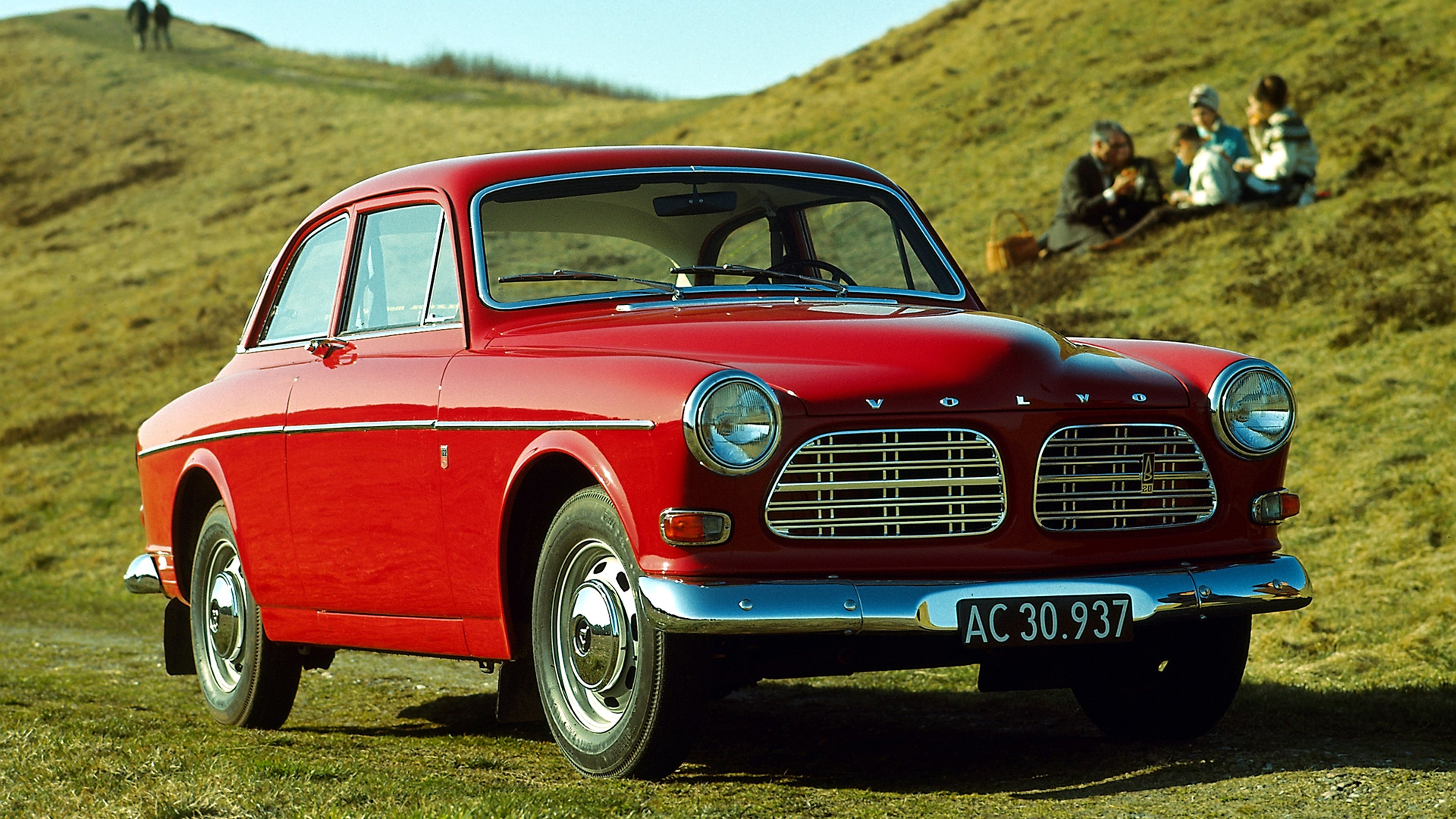 Classica Volvo Amazon di colore rosso, con griglia cromata e fari rotondi, parcheggiata su una collina erbosa, con persone che si rilassano sullo sfondo sotto un cielo azzurro e limpido.