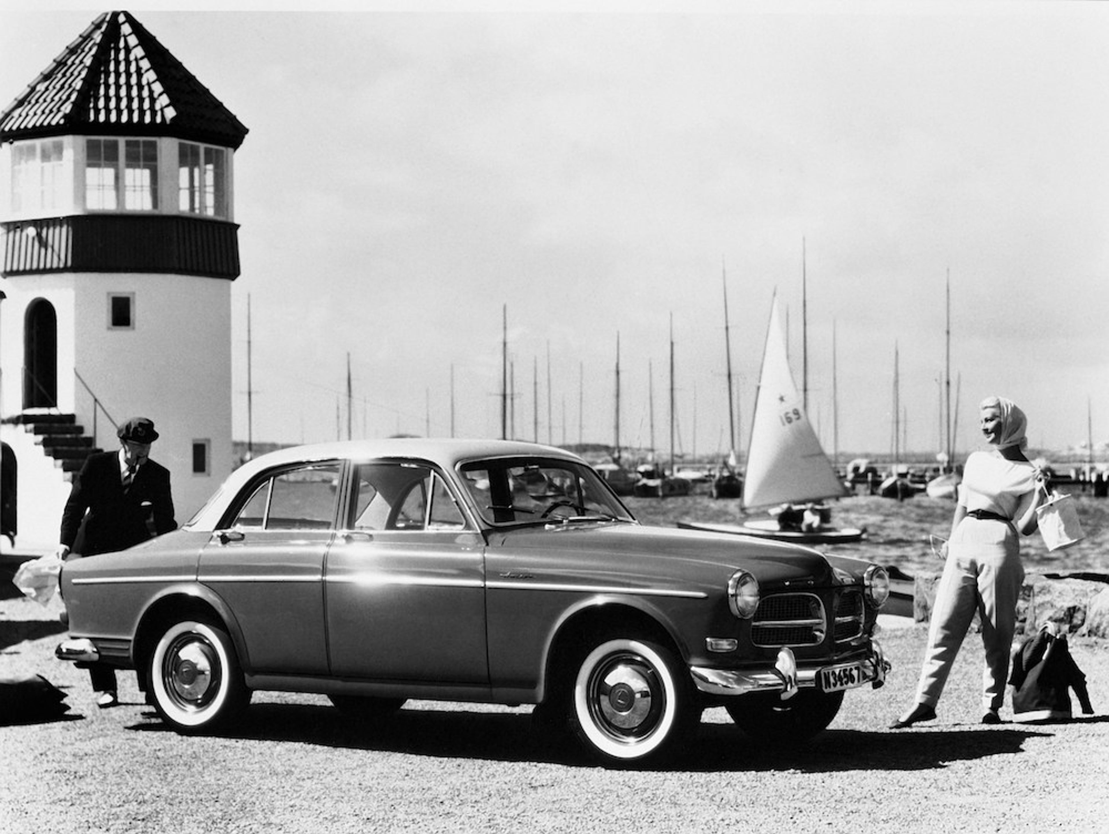 Der Volvo 122 Amazon als Schwarz-Weiss Aufnahme. Eine Frau steht vor ihm und ein Mann dahinter. Sie befinden sich am Hafen neben einem Leuchtturm.