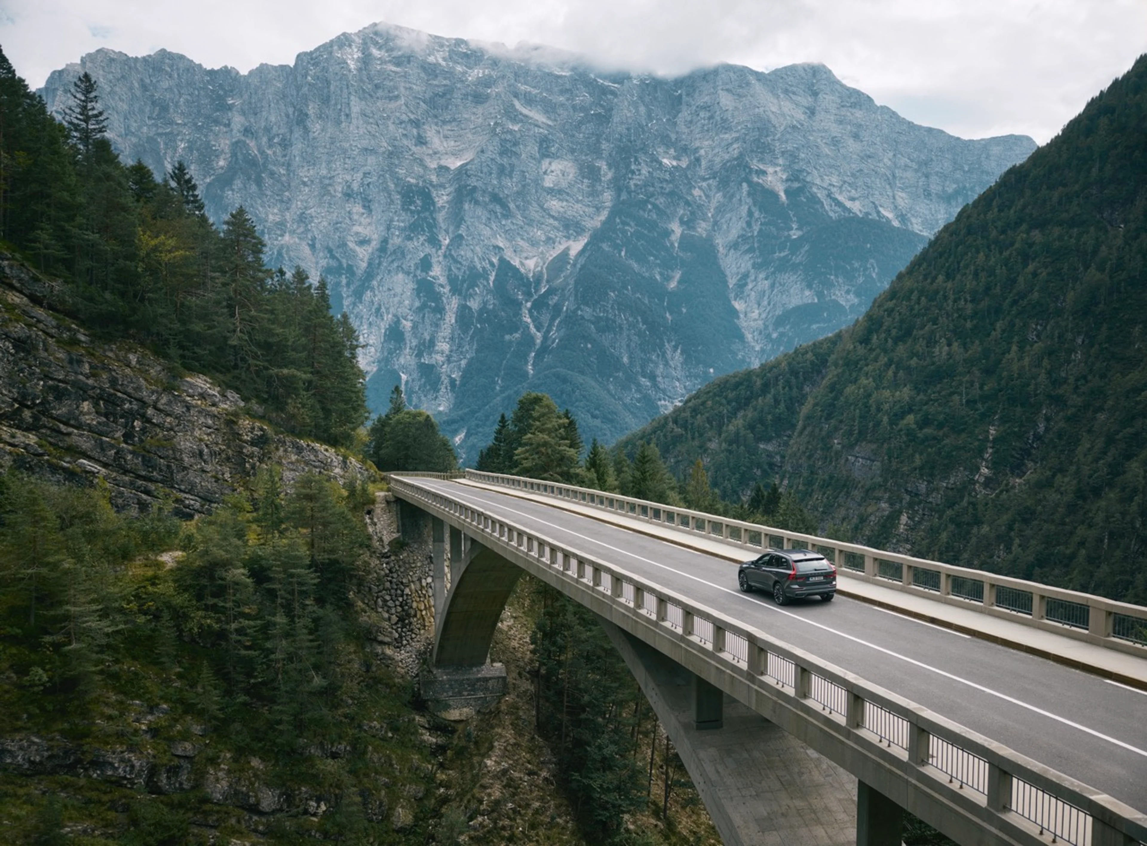 A Volvo XC60 crosses a bridge over a forested valley with rugged mountains in the background.