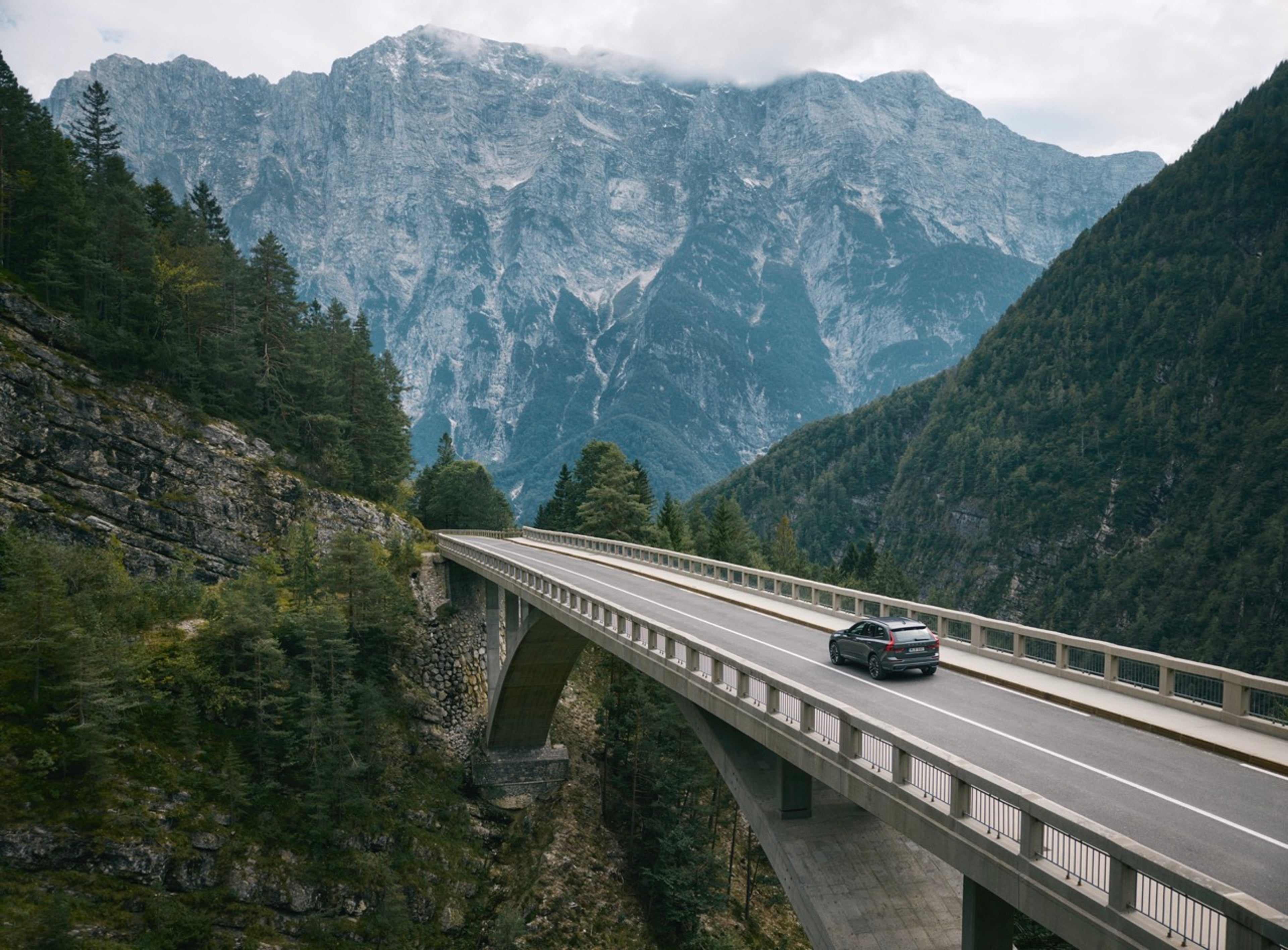 A Volvo XC60 crosses a bridge over a forested valley with rugged mountains in the background.