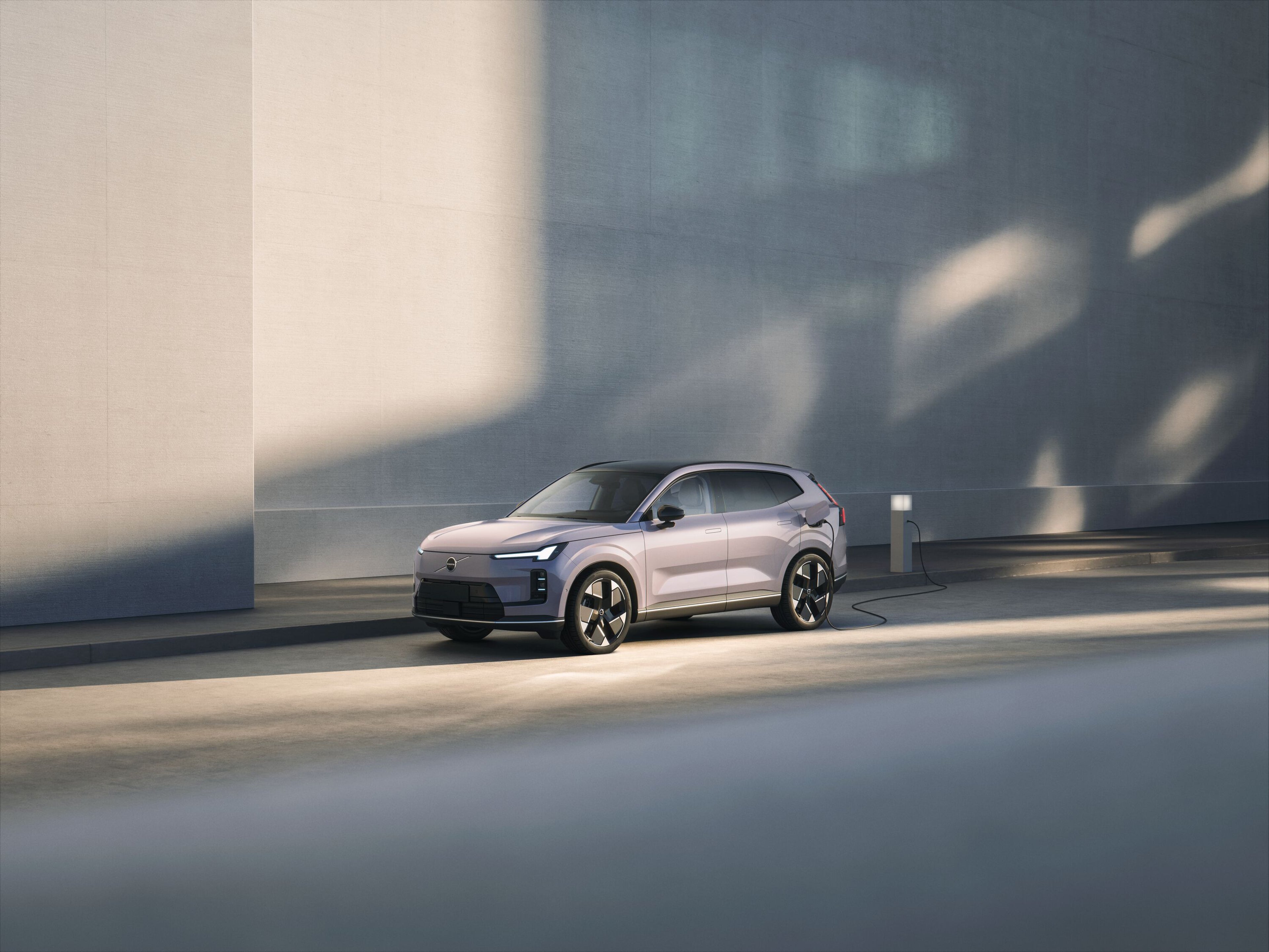 Elektryczne Volvo XC70 zaparkowane na nasłonecznionej ulicy, podłączone do stacji ładowania obok wysokiego muru.