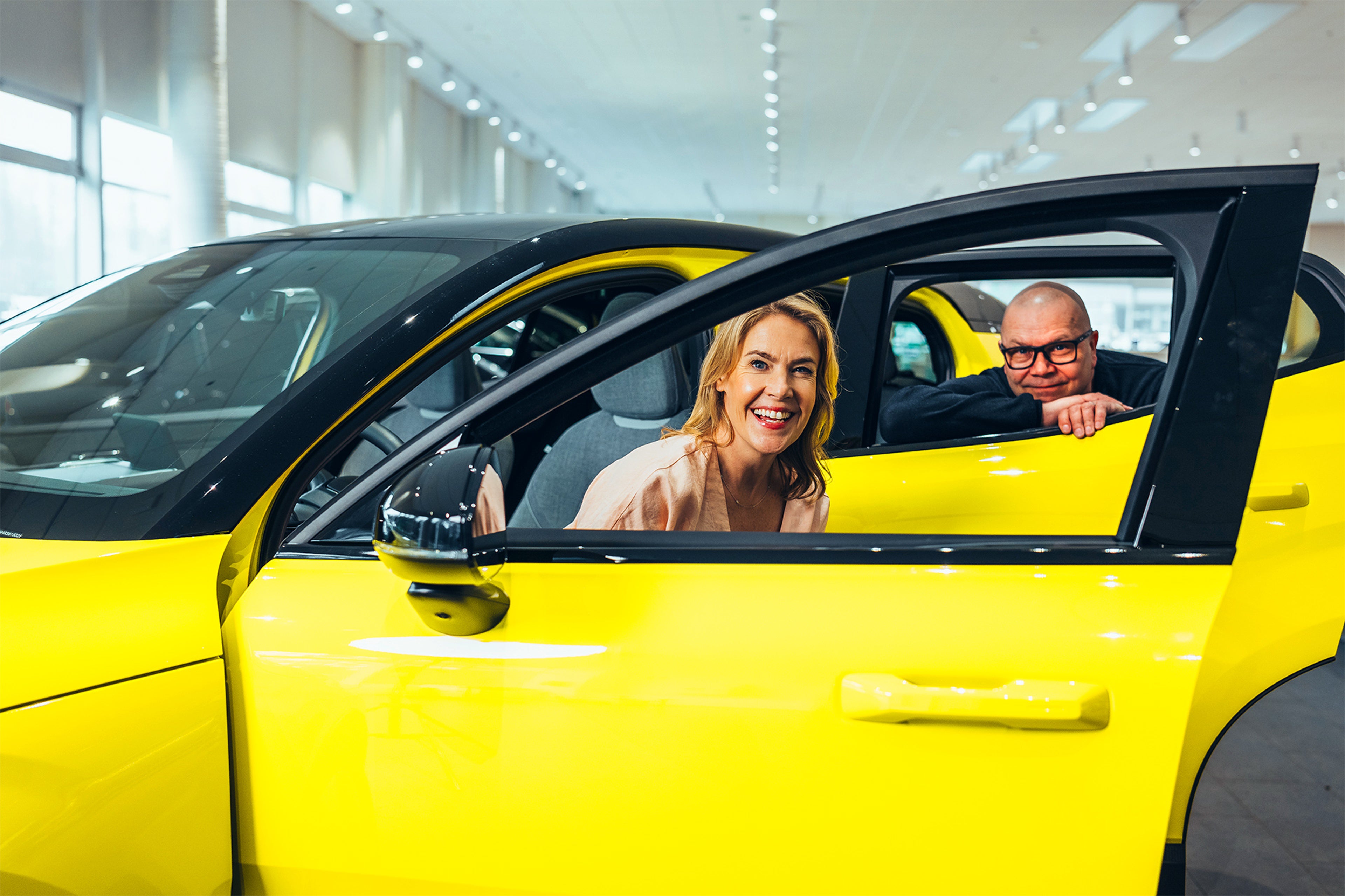Ella Kanninen ja Ari Parkkonen kurkistavat Moss Yellow -värisen Volvo EX30 täyssähköauton ikkunoista.