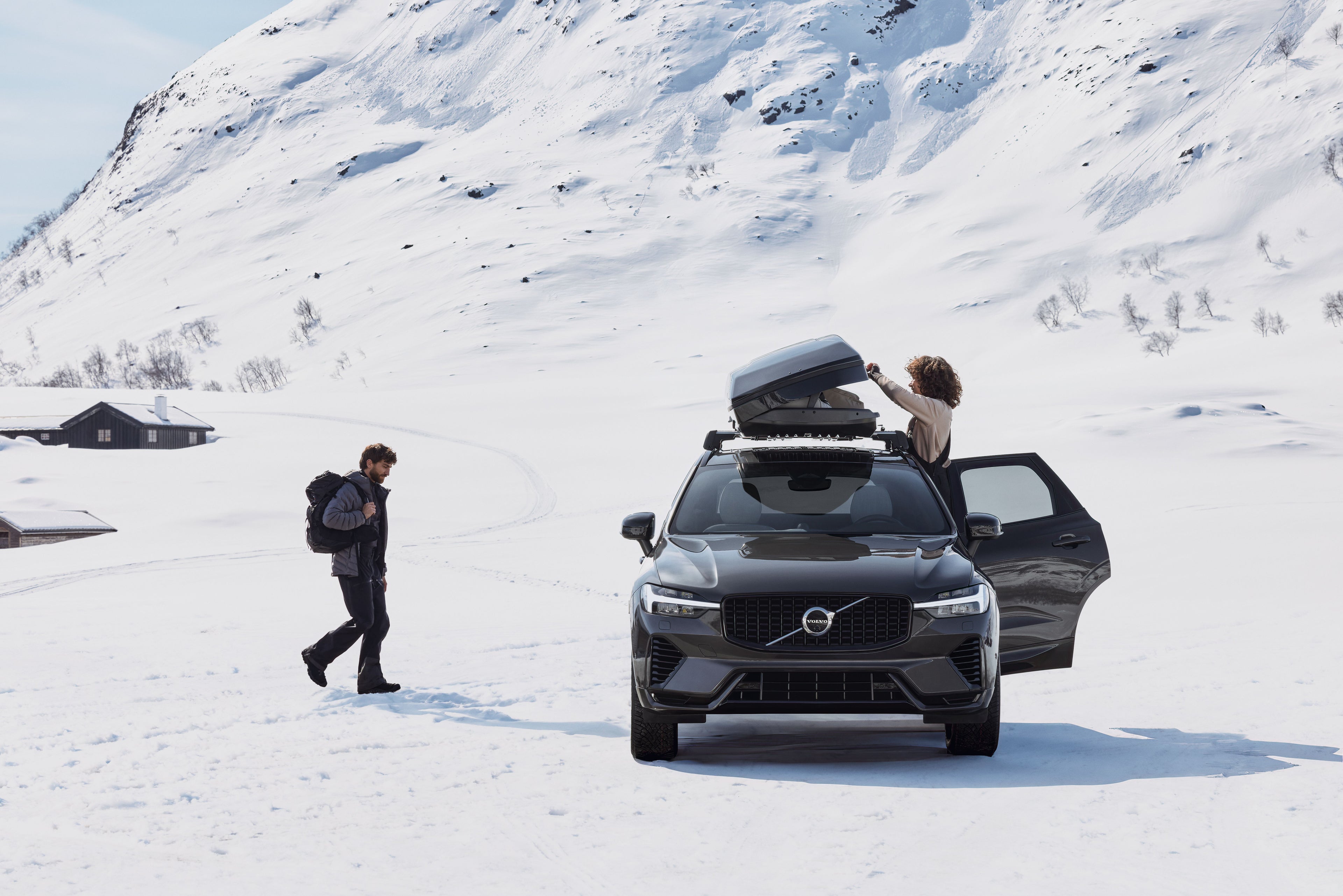 Una mujer abre el cofre de techo de un Volvo XC60 negro aparcado junto a una montaña nevada, mientras un hombre camina hacia ella