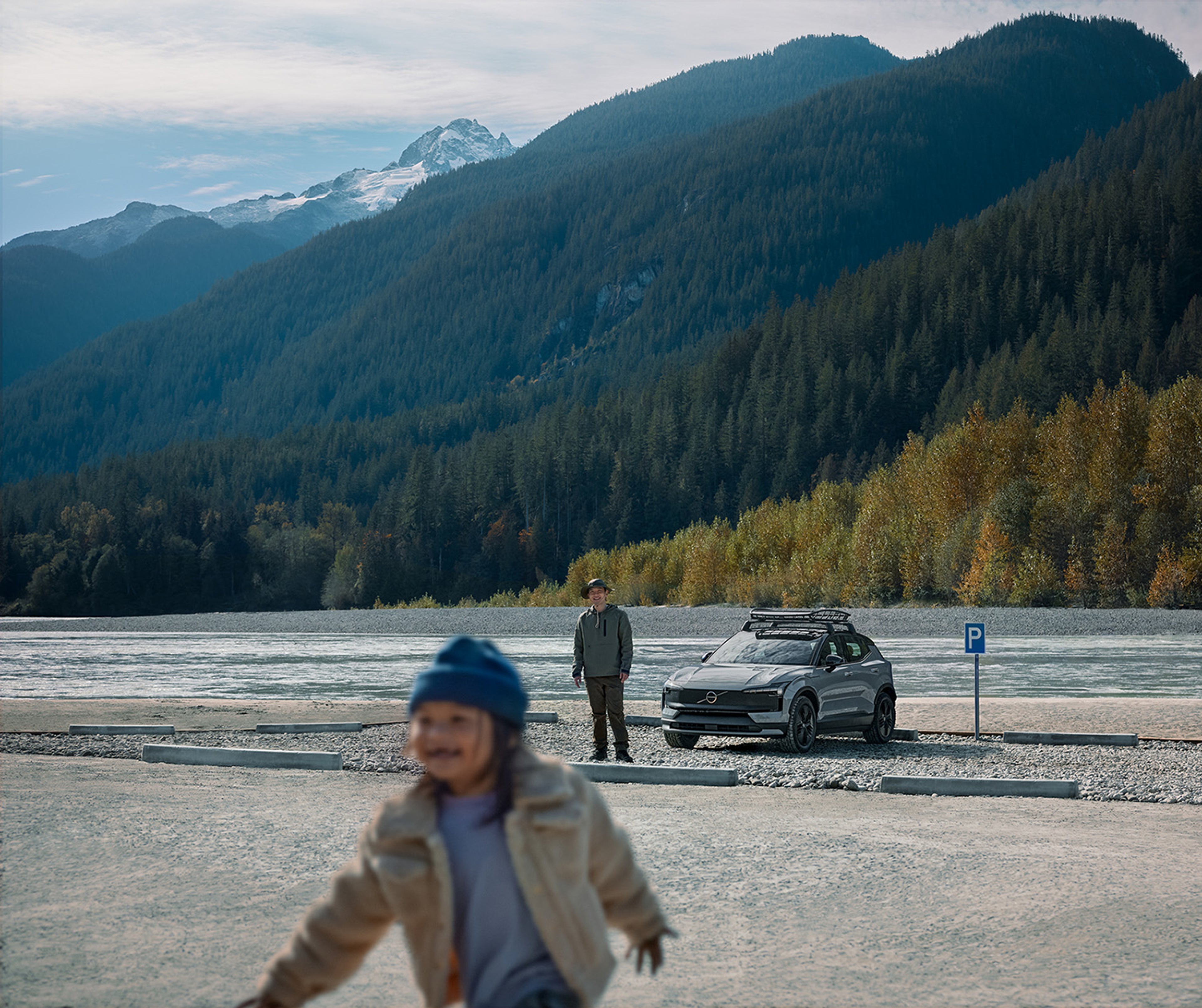 A Vapour Grey Volvo EX30CC with a roof rack is parked by a river with mountains in the background. A person in outdoor clothing stands beside the car, while a child in a blue hat and beige jacket moves in the foreground.