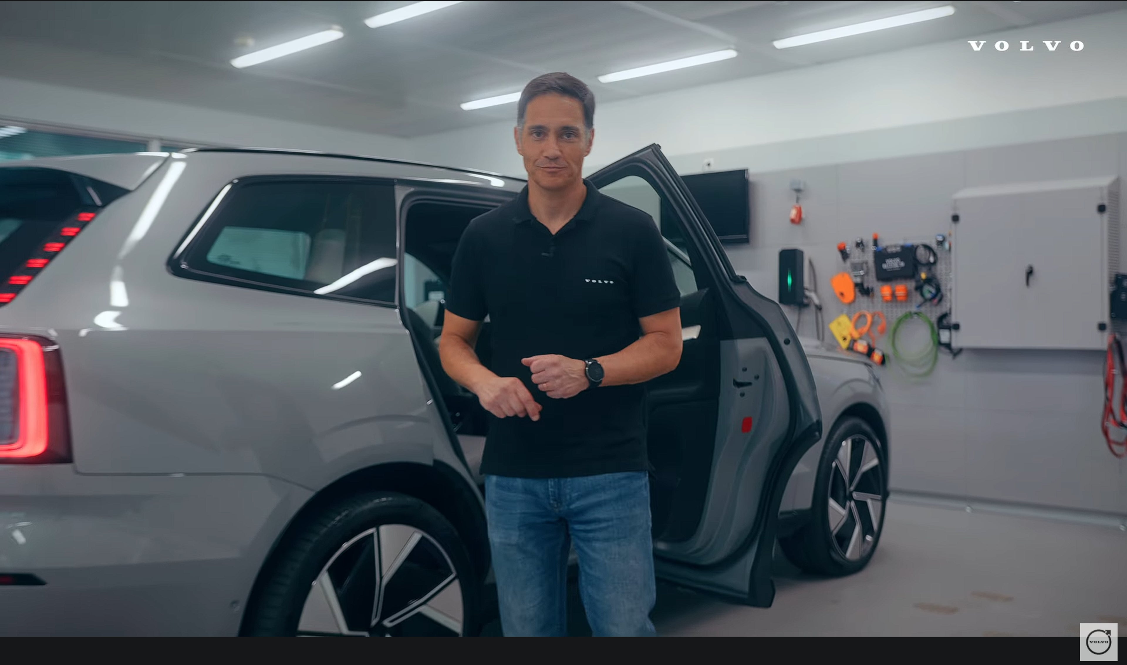 Expert Volvo numa oficina moderna, ao lado de um Volvo EX90 cinzento com a porta traseira aberta, durante uma explicação técnica sobre o veículo elétrico.