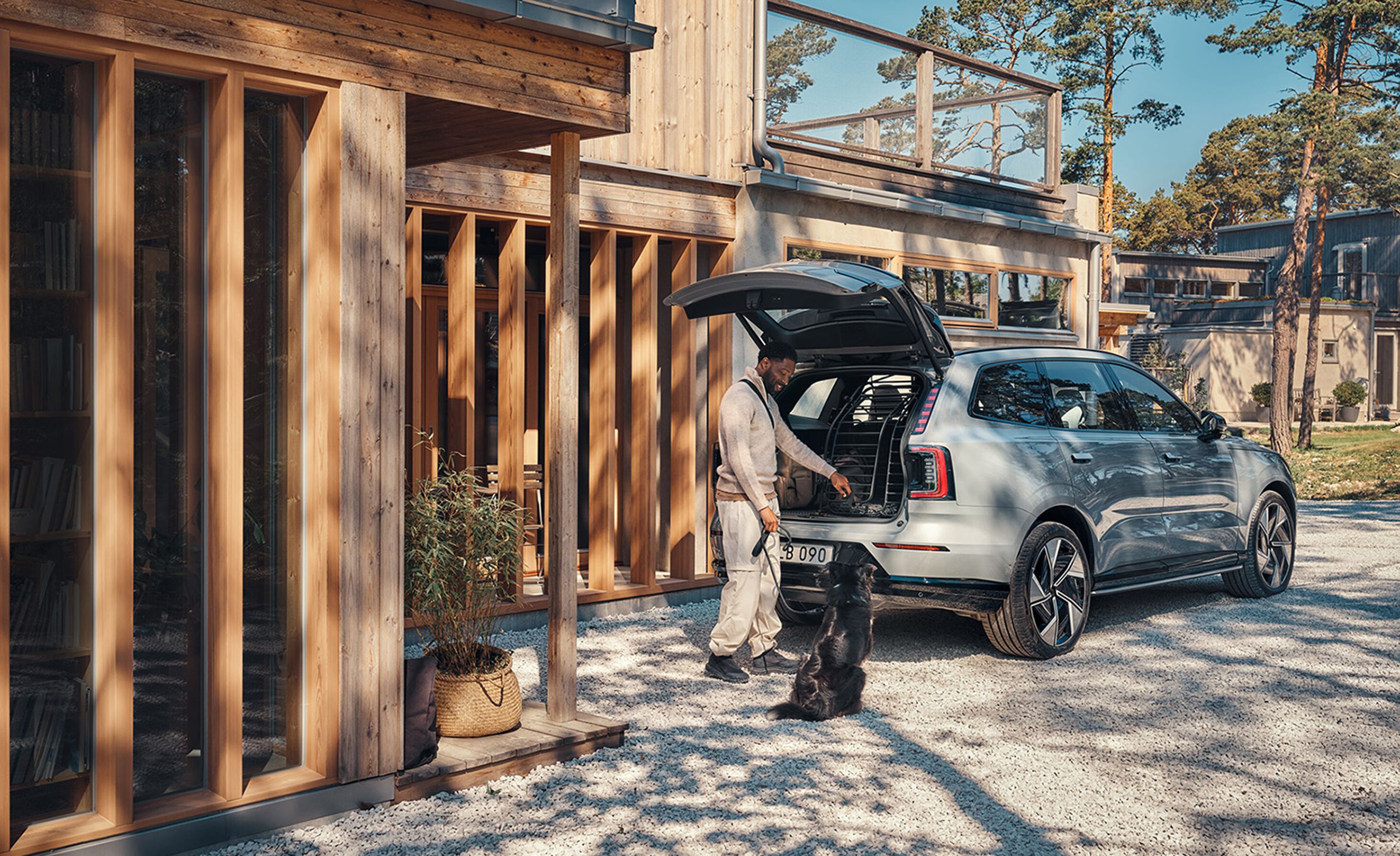 رجل يشجع كلبه على القفز في صندوق سيارته، خارج منزل في يوم مشمس.