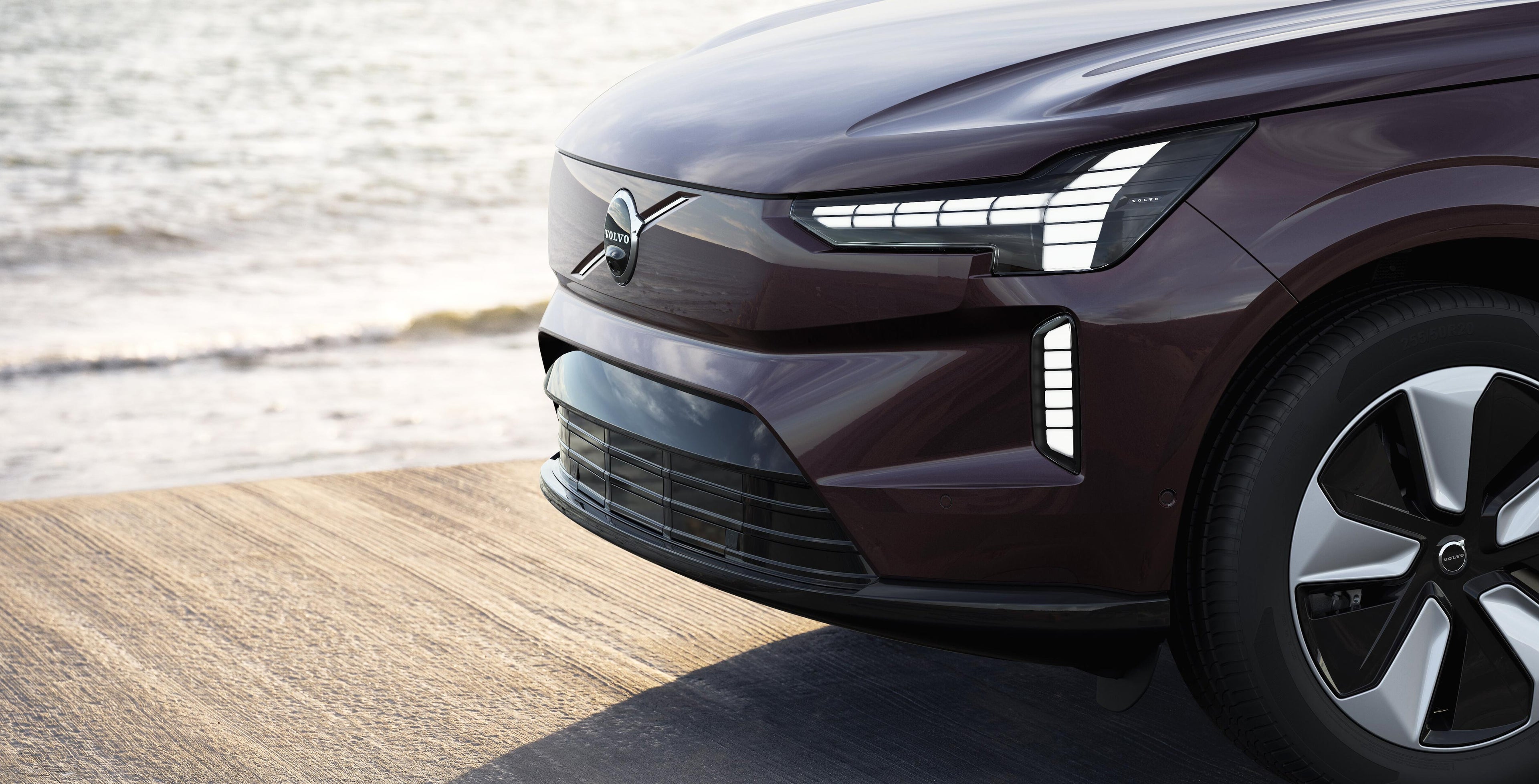 Veden ääreen betonilaiturille pysäköidyn Mulberry Red värisen Volvo EX90 Plus auton keula.