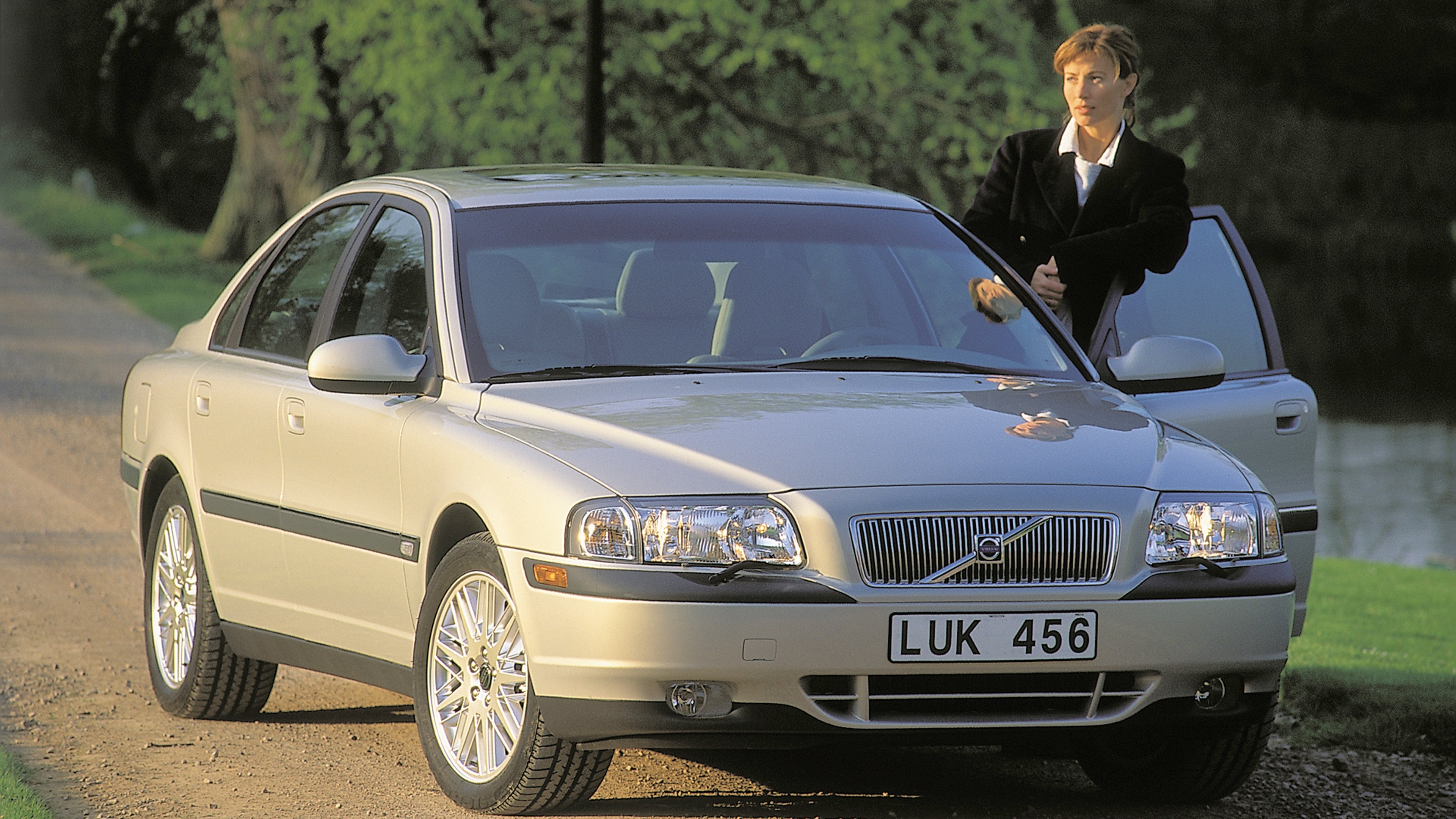 Роскошный седан Volvo S80 Gen 1 серебристого цвета припаркован на живописной грунтовой дороге в окружении зелени, рядом с открытой водительской дверью стоит человек.