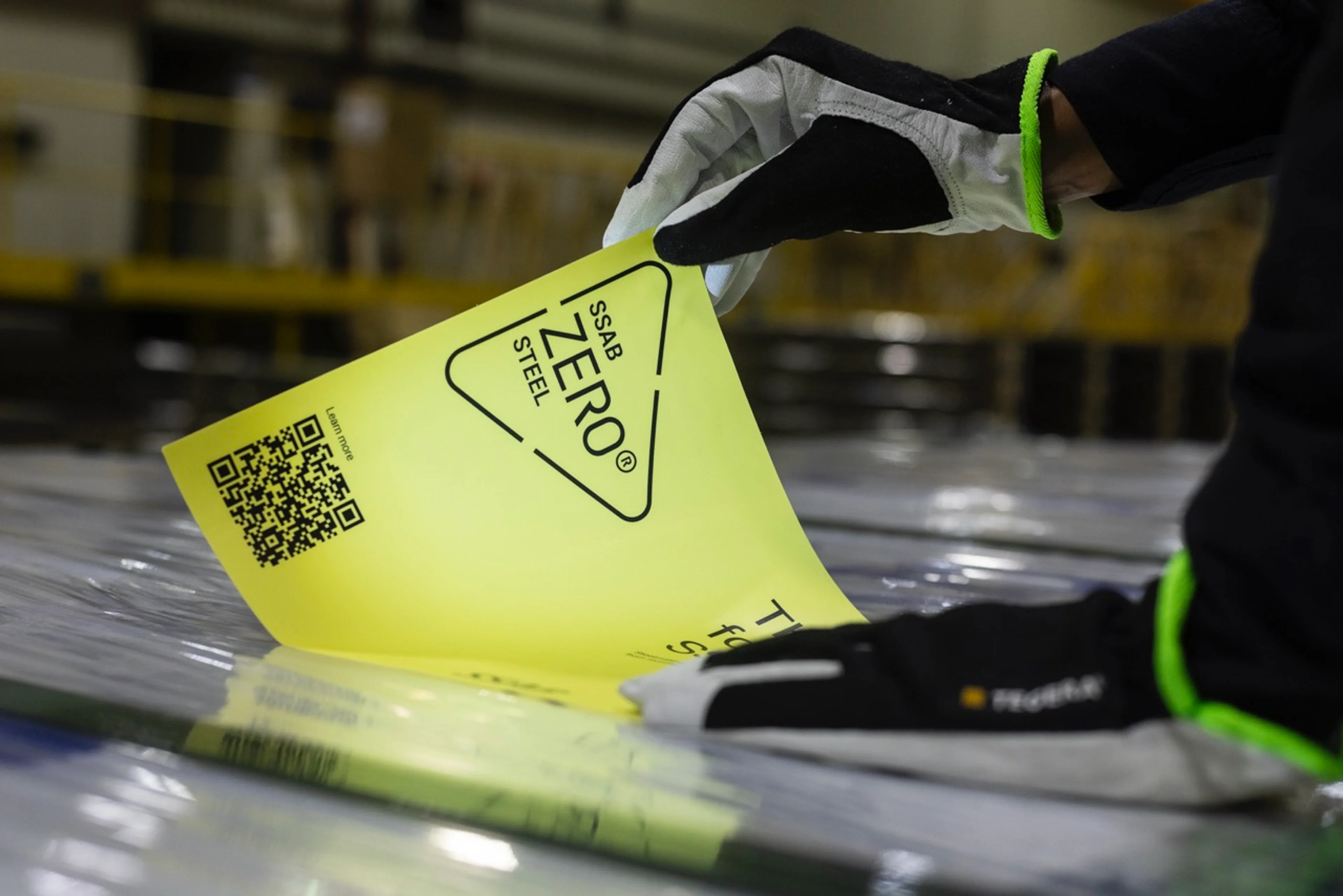 Un par de manos enguantadas despegan una etiqueta amarilla con "SSAB ZERO® STEEL" impresa en ella, revelando una superficie de acero debajo.