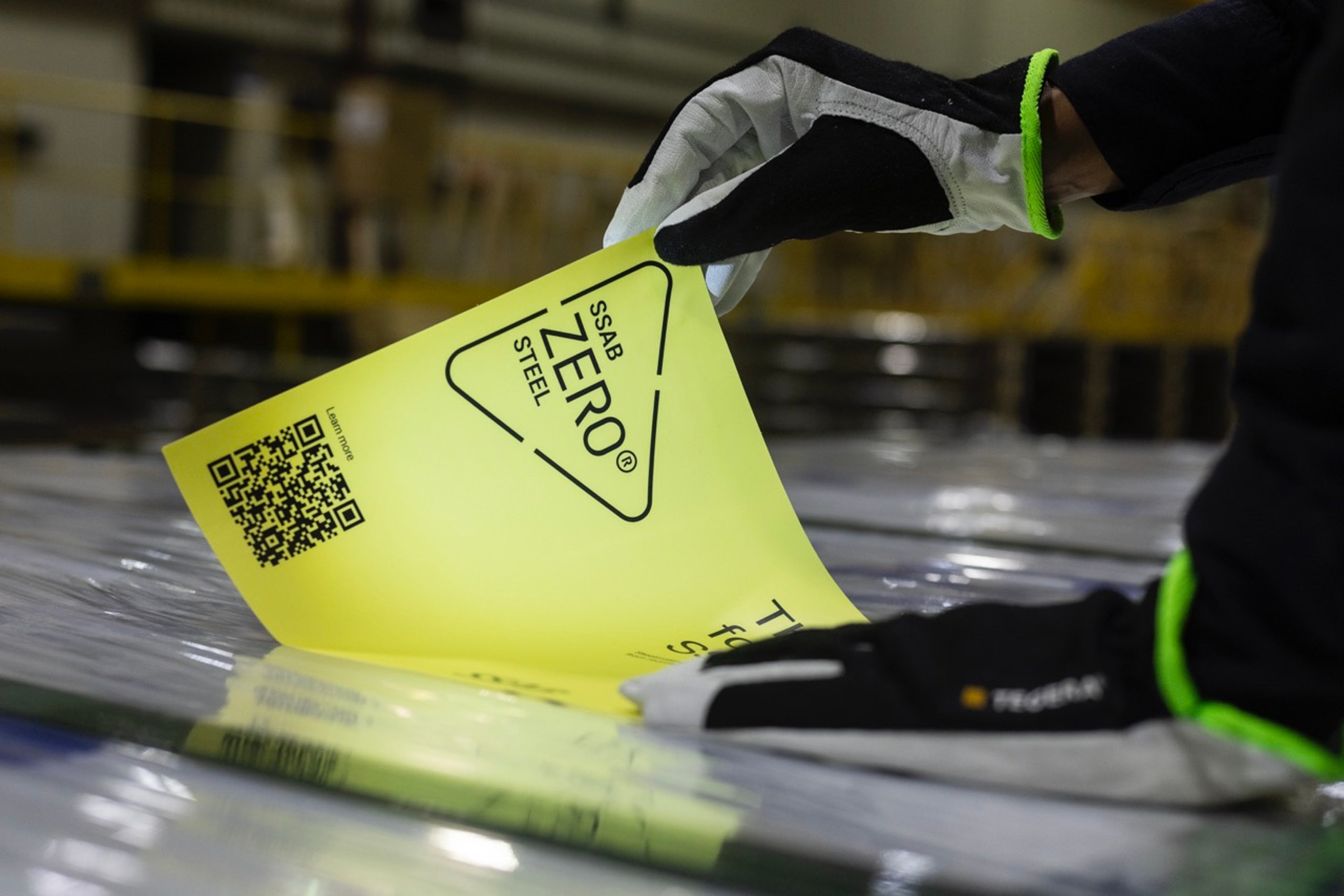 تنزع يدان ترتديان القفازات ملصقاً أصفر مطبوعاً عليه عبارة "SSAB ZERO® STEEL"، مما يكشف عن سطح فولاذي تحته.